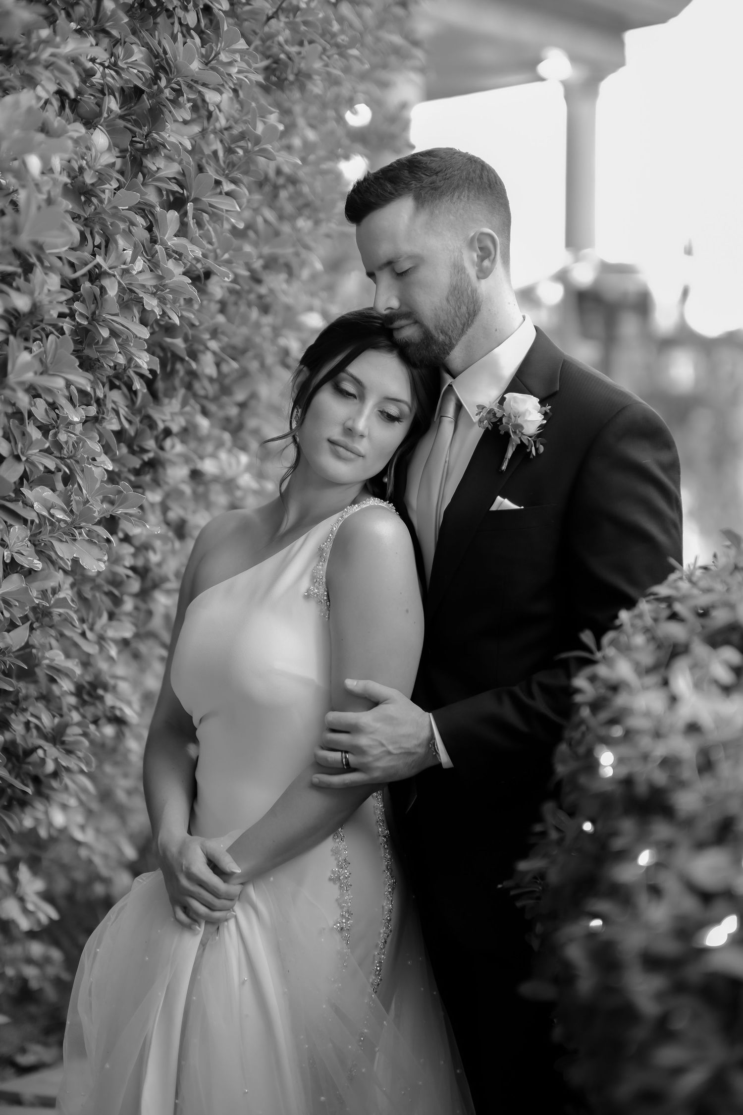 Romantic black and white portrait of newlyweds embracing in a garden setting with soft lighting.