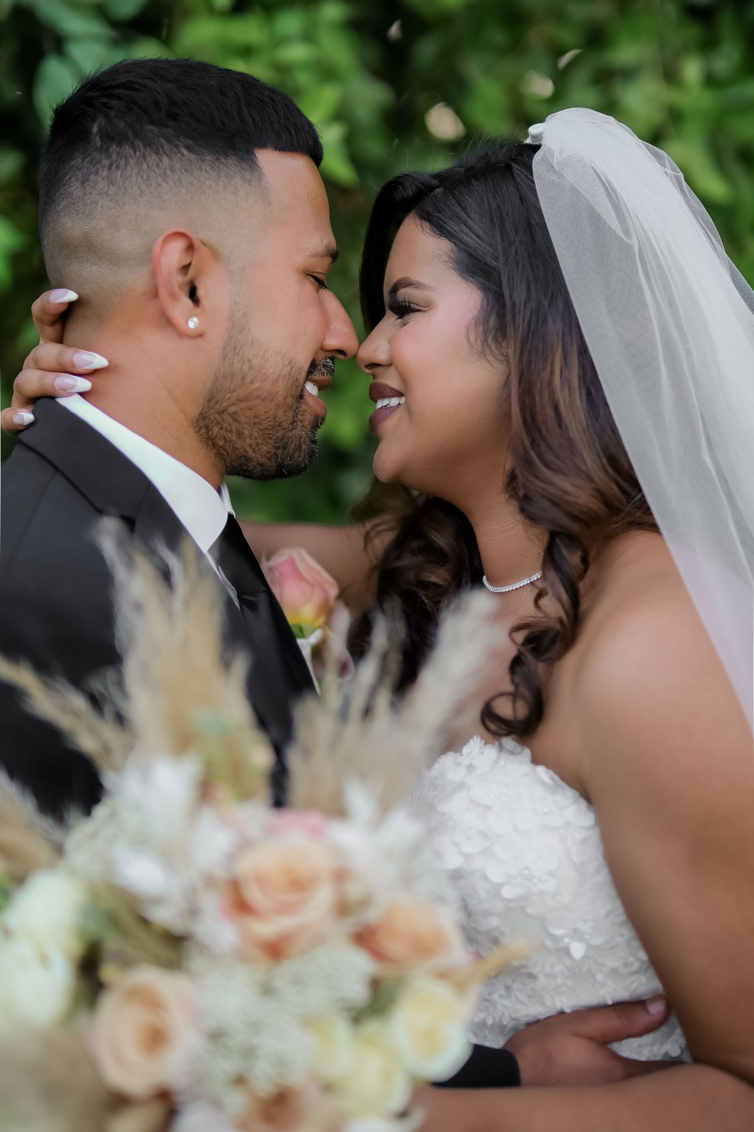 A bride and groom share a romantic moment, smiling at each other with a blurred floral bouquet in the foreground.