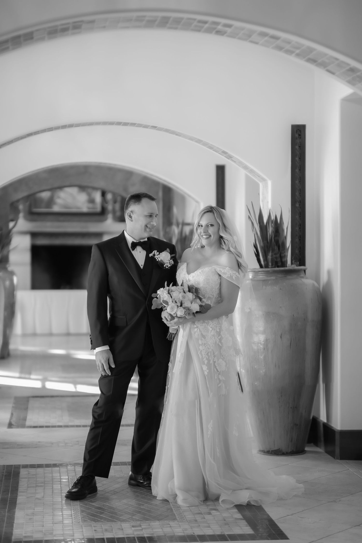 A wedding couple stands in an elegant hallway with arched ceilings and a large decorative vase.