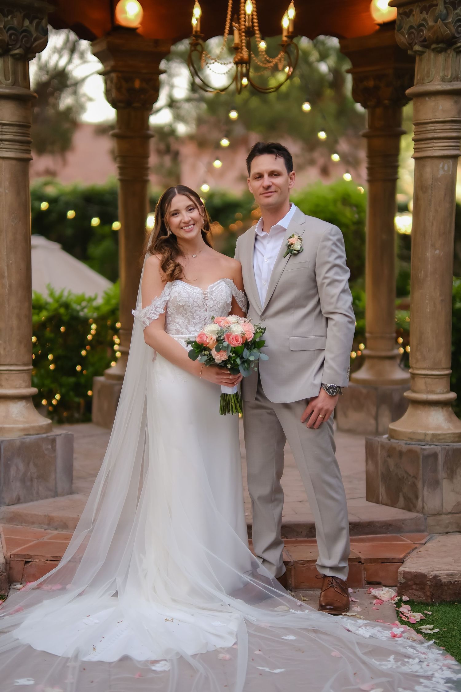 Bride in white gown holding pink bouquet stands with groom in tan suit under elegant gazebo with string lights.