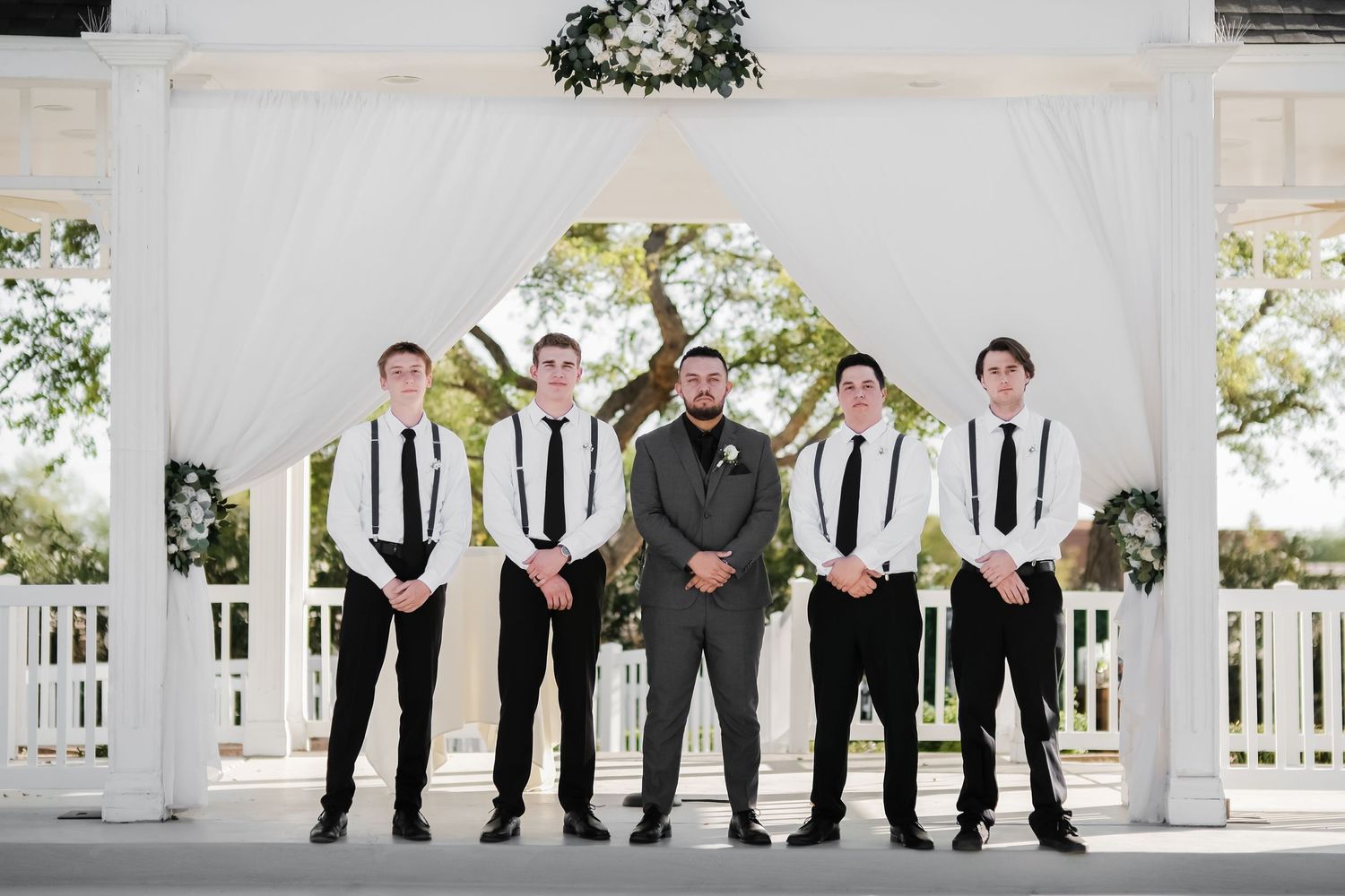 Groomsmen wearing black and white suits with striped ties pose together under an elegant white gazebo.