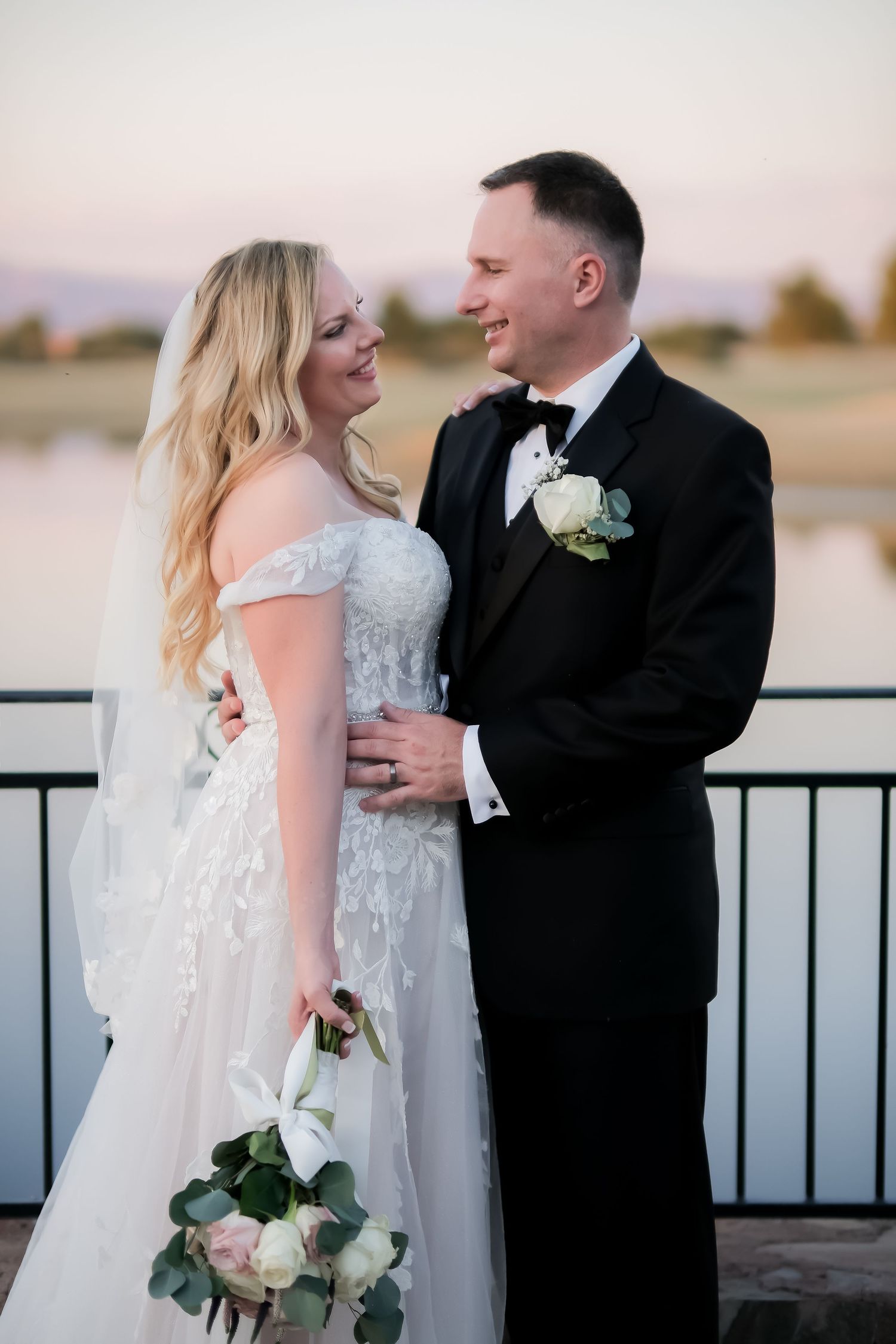A bride in a white lace gown and groom in a black tuxedo share a romantic moment by a lakeside at sunset.