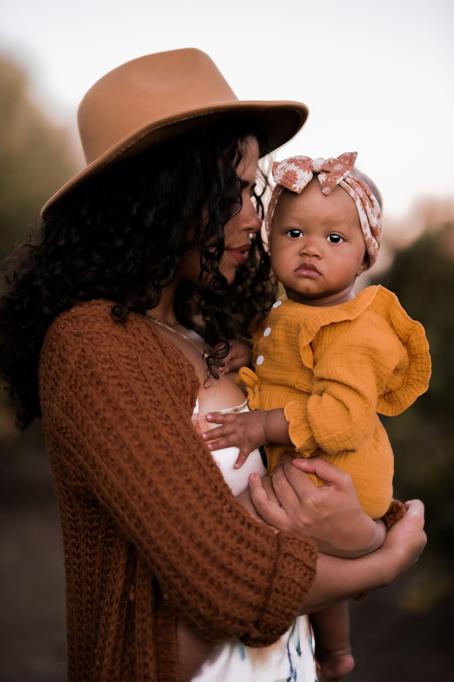 Portrait of parent wearing brown sweater and hat holding baby in matching mustard outfit.