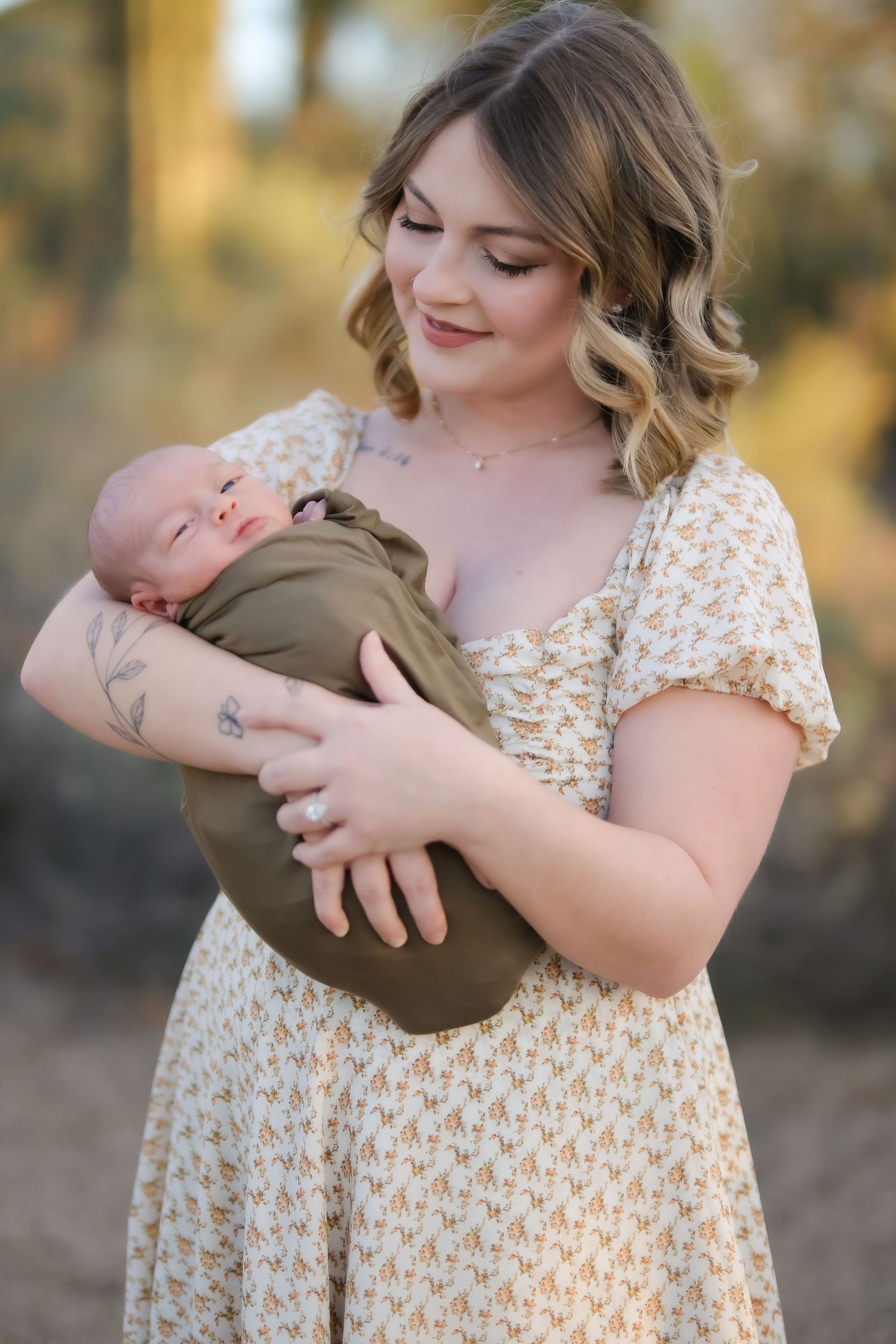 Mother in floral dress tenderly holds sleeping newborn wrapped in olive swaddle.