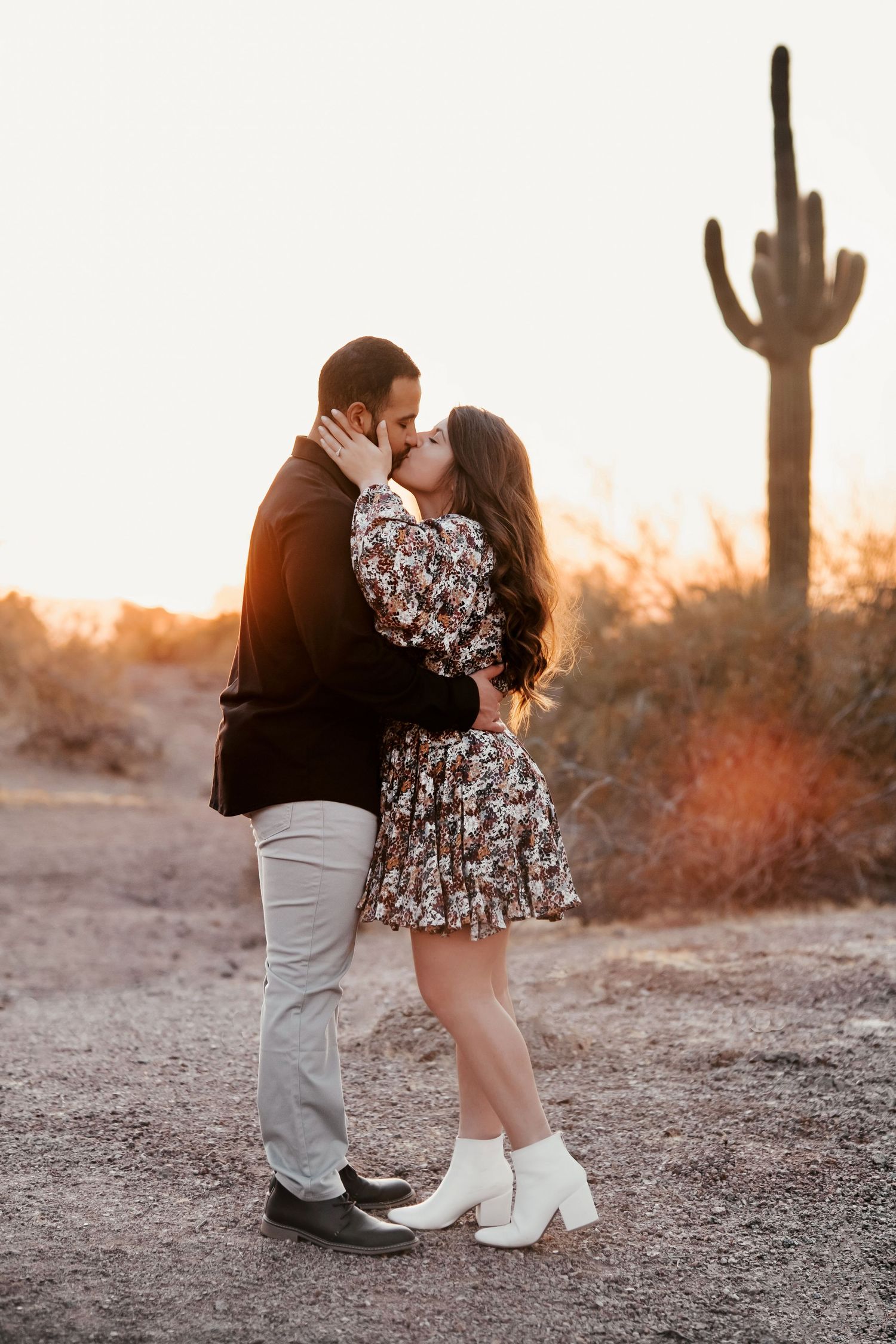 Romantic desert engagement photo with couple embracing near a saguaro cactus at sunset.
