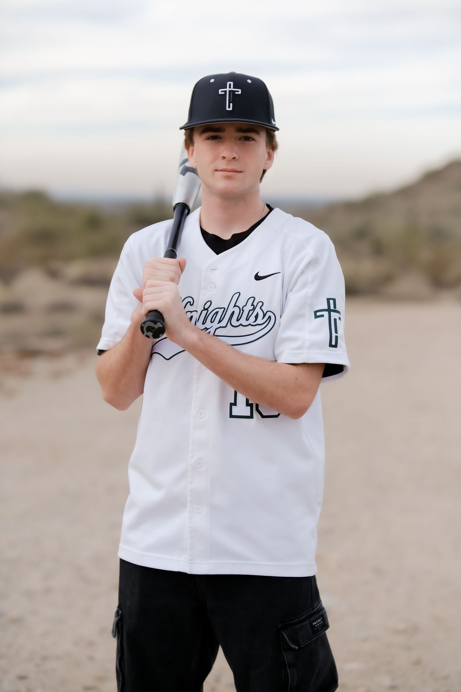 Baseball player in white uniform and cap with cross emblem poses with bat in desert setting.