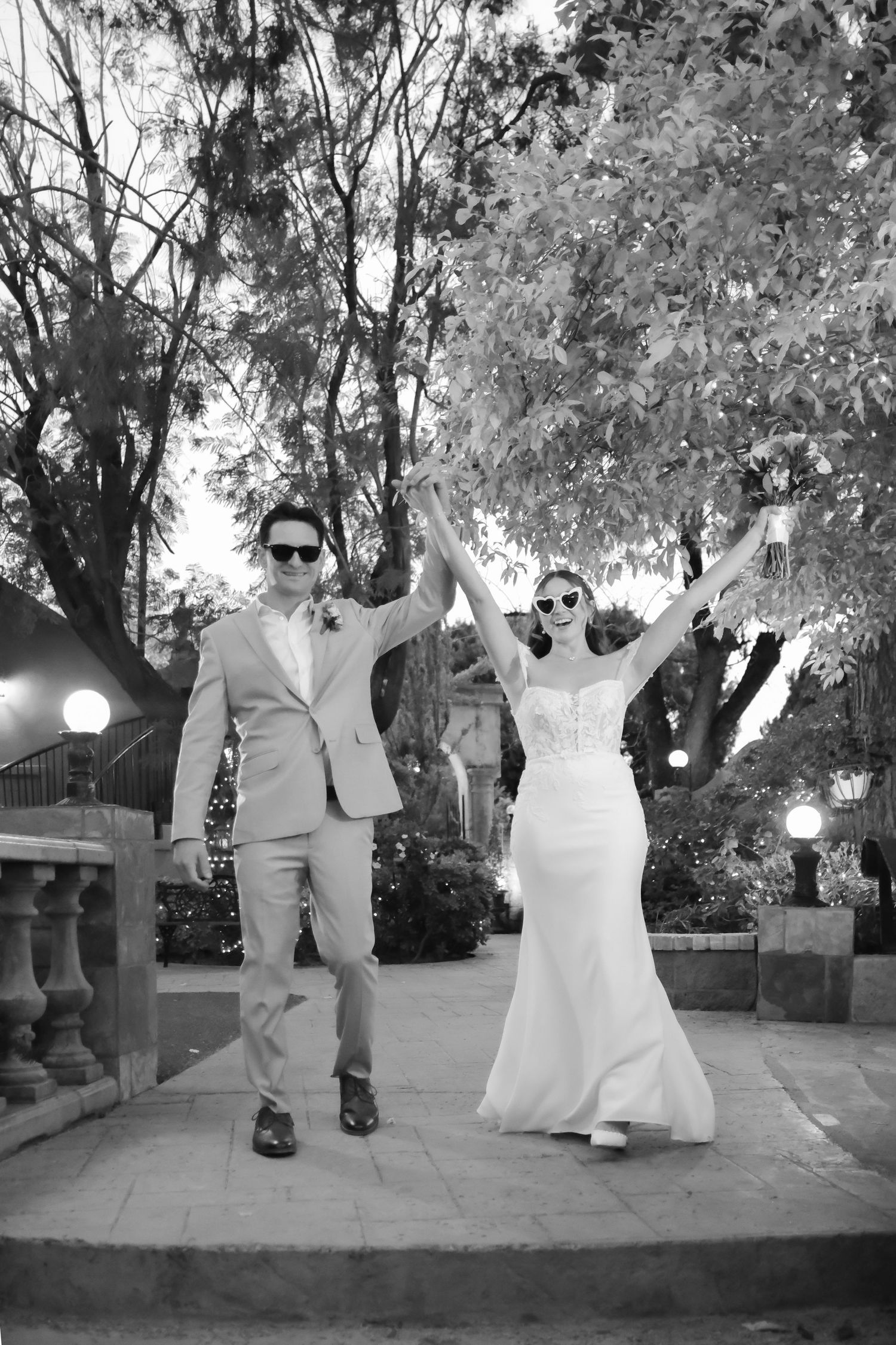 A joyful wedding couple in formal attire celebrating with raised arms in black and white.