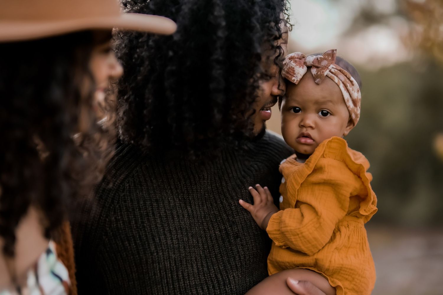 Baby in mustard yellow outfit looking curiously while being held in outdoor portrait.