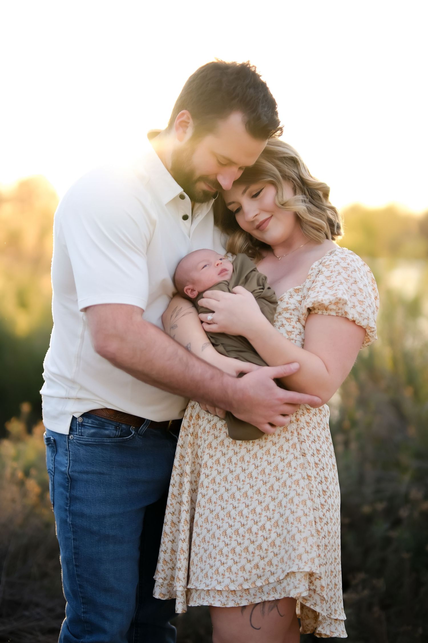 New parents embrace while holding their newborn baby during sunset photoshoot.