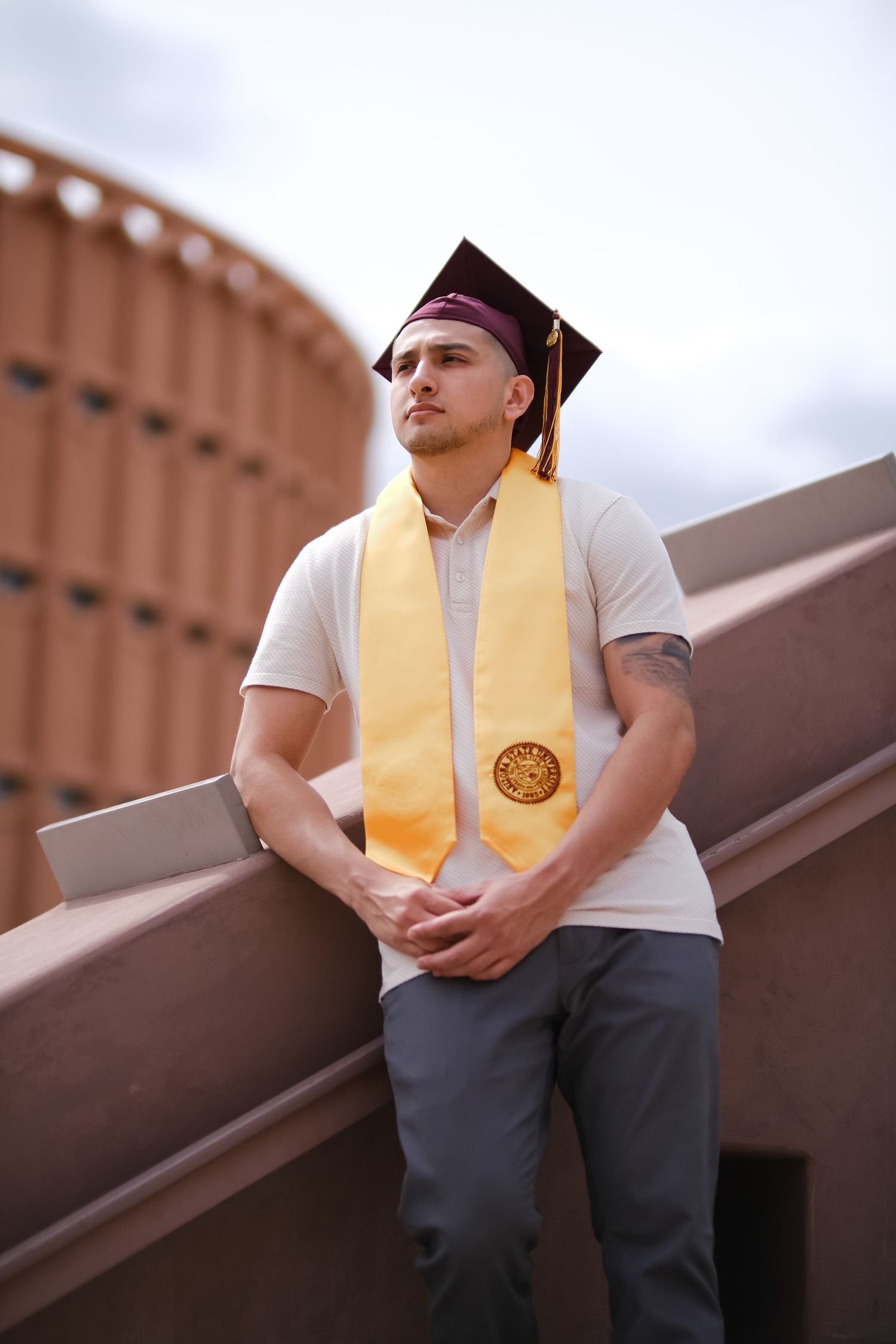 Graduate in burgundy cap and gold stole stands thoughtfully on stairs with industrial building in background.
