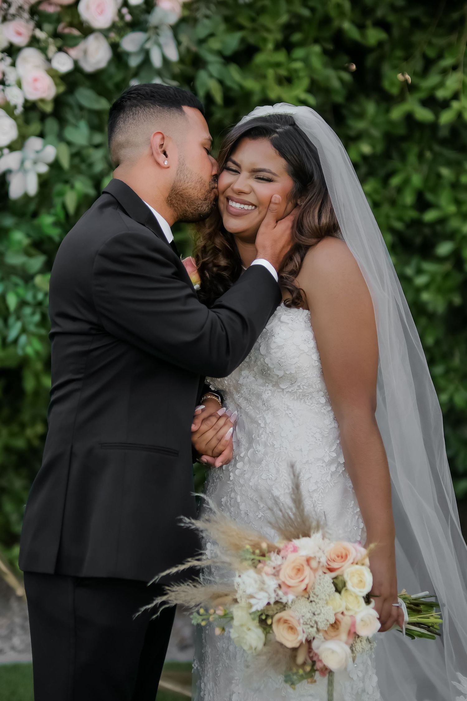 Groom in black tuxedo kisses smiling bride in white lace gown holding pastel bouquet outdoors.