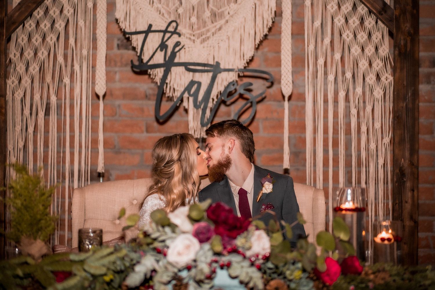 Newlyweds share a romantic kiss at sweetheart table adorned with lush floral arrangements beneath a macrame backdrop.