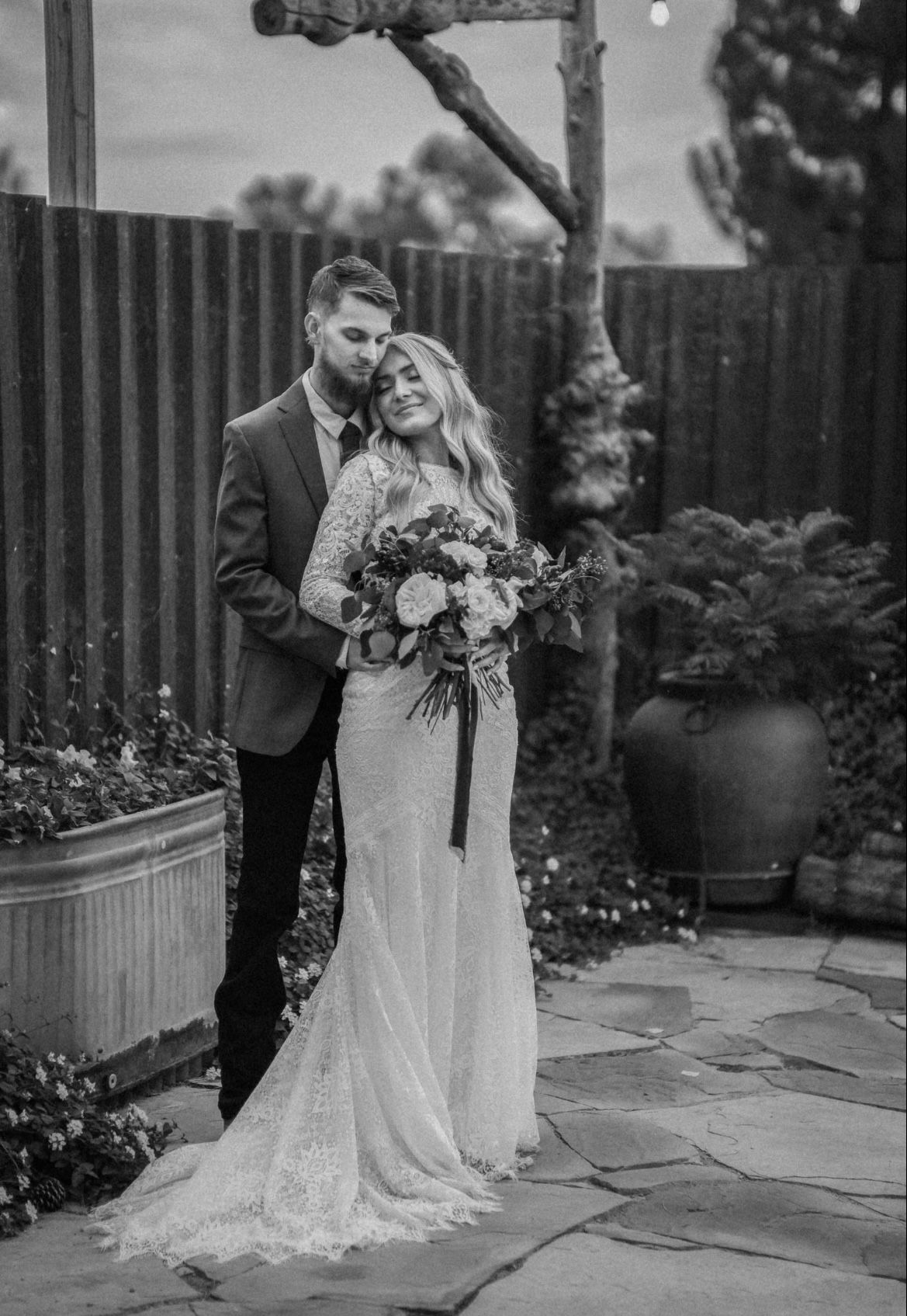 Black and white portrait of couple embracing in garden setting with stone patio and planters.