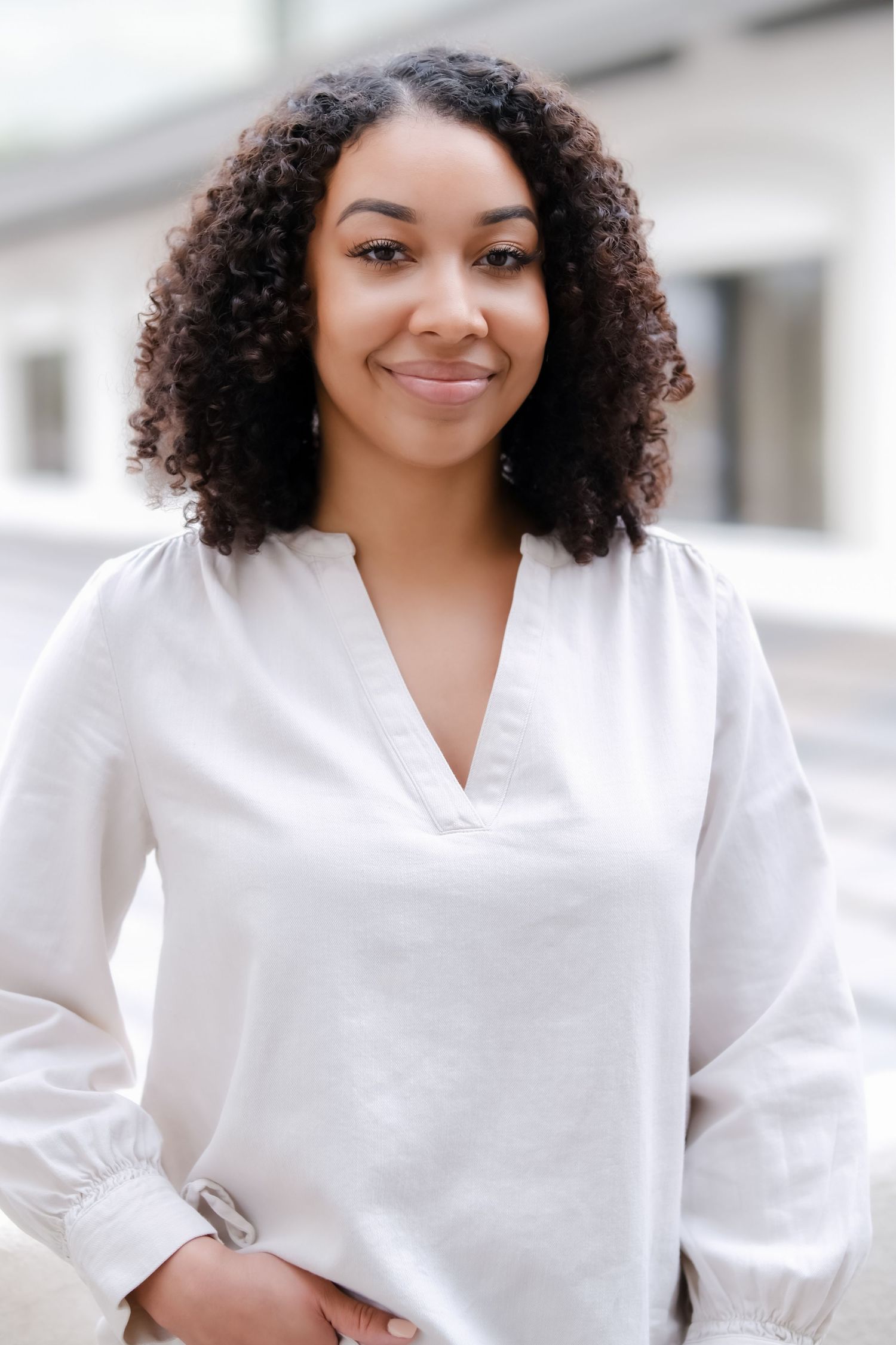 Professional headshot of woman with curly hair wearing white blouse against light background.