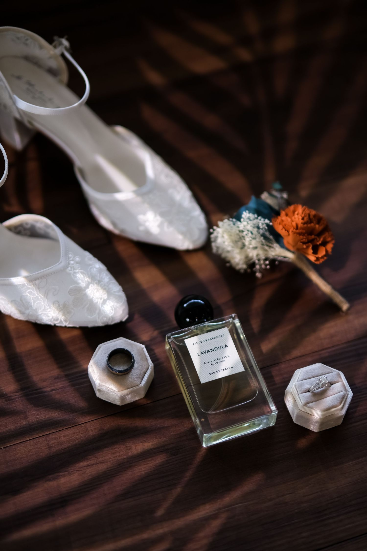 Wedding details including white lace shoes, perfume bottle, and floral boutonniere on dark wooden surface.