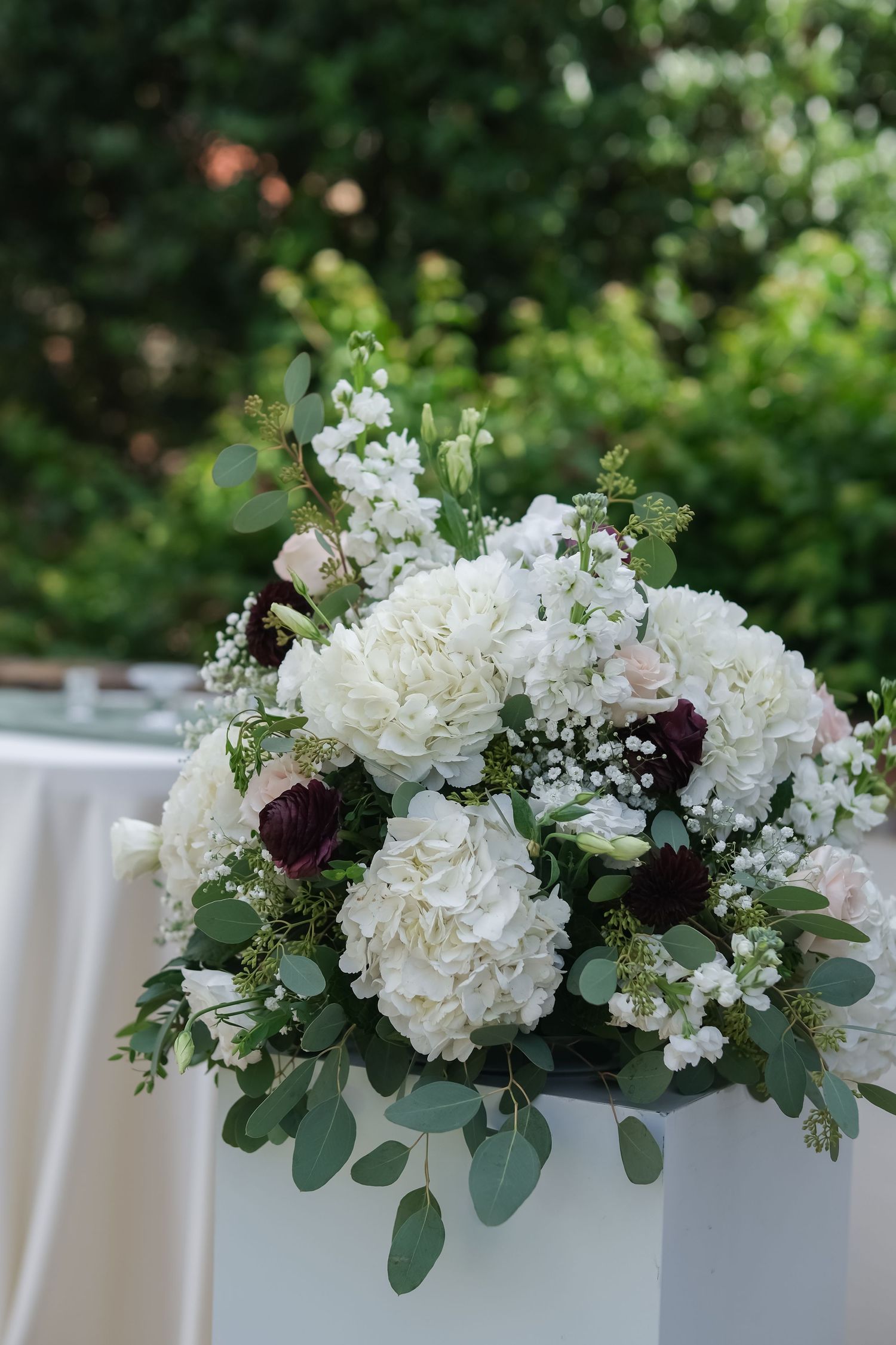 Elegant wedding floral arrangement with white hydrangeas, burgundy roses, baby's breath, and eucalyptus on a white pedestal.