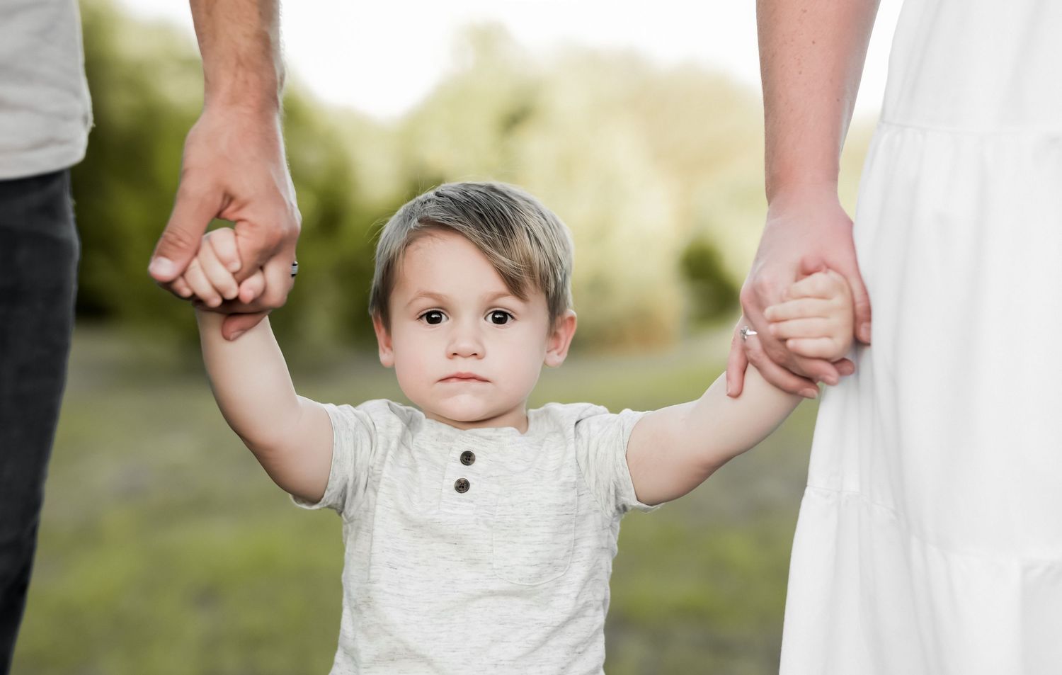 Small toddler holding both parents' hands while standing between them outdoors.