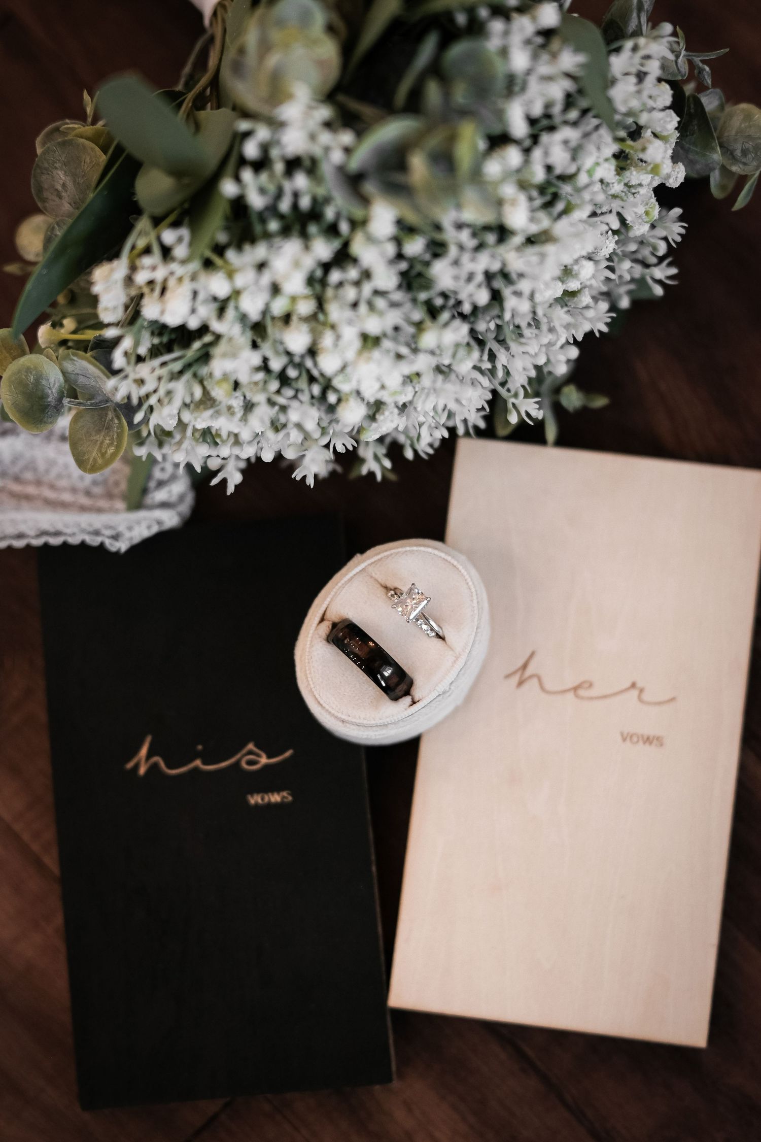 Wedding vow books labeled 'his' and 'her' with ring box and white floral arrangement.