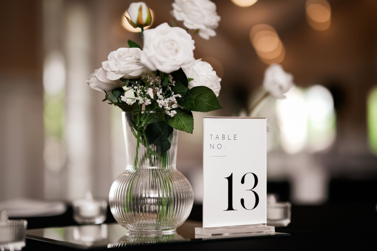 Table number 13 card displayed next to a glass vase of white roses with blurred background.