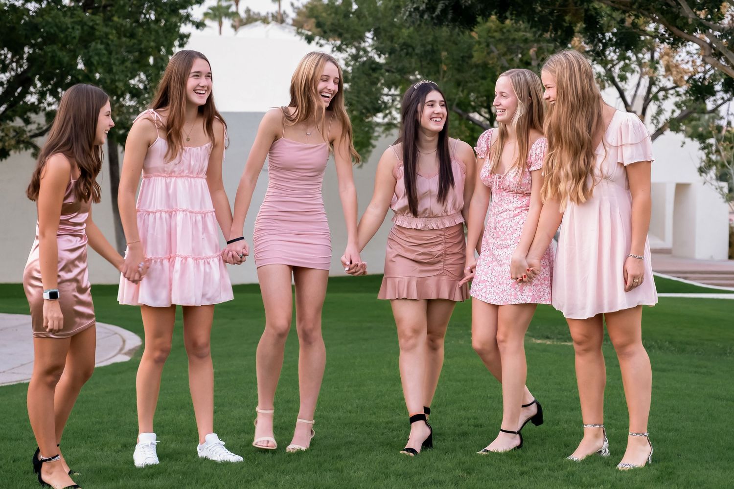 A group wearing coordinated pink dresses and white shoes poses together on a green lawn.