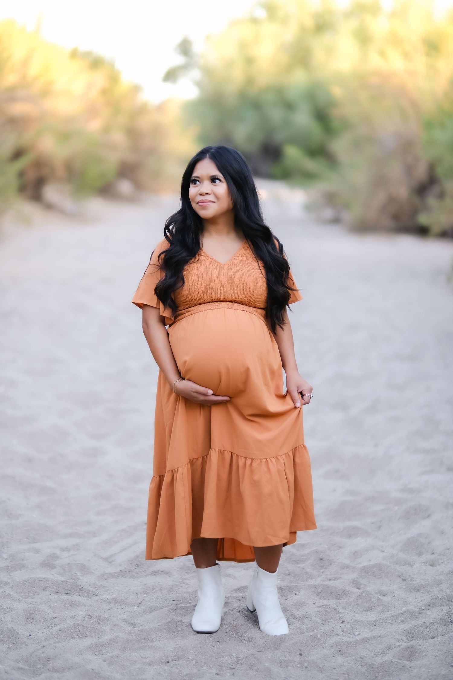 Expectant mother in rust-colored dress poses in desert setting.