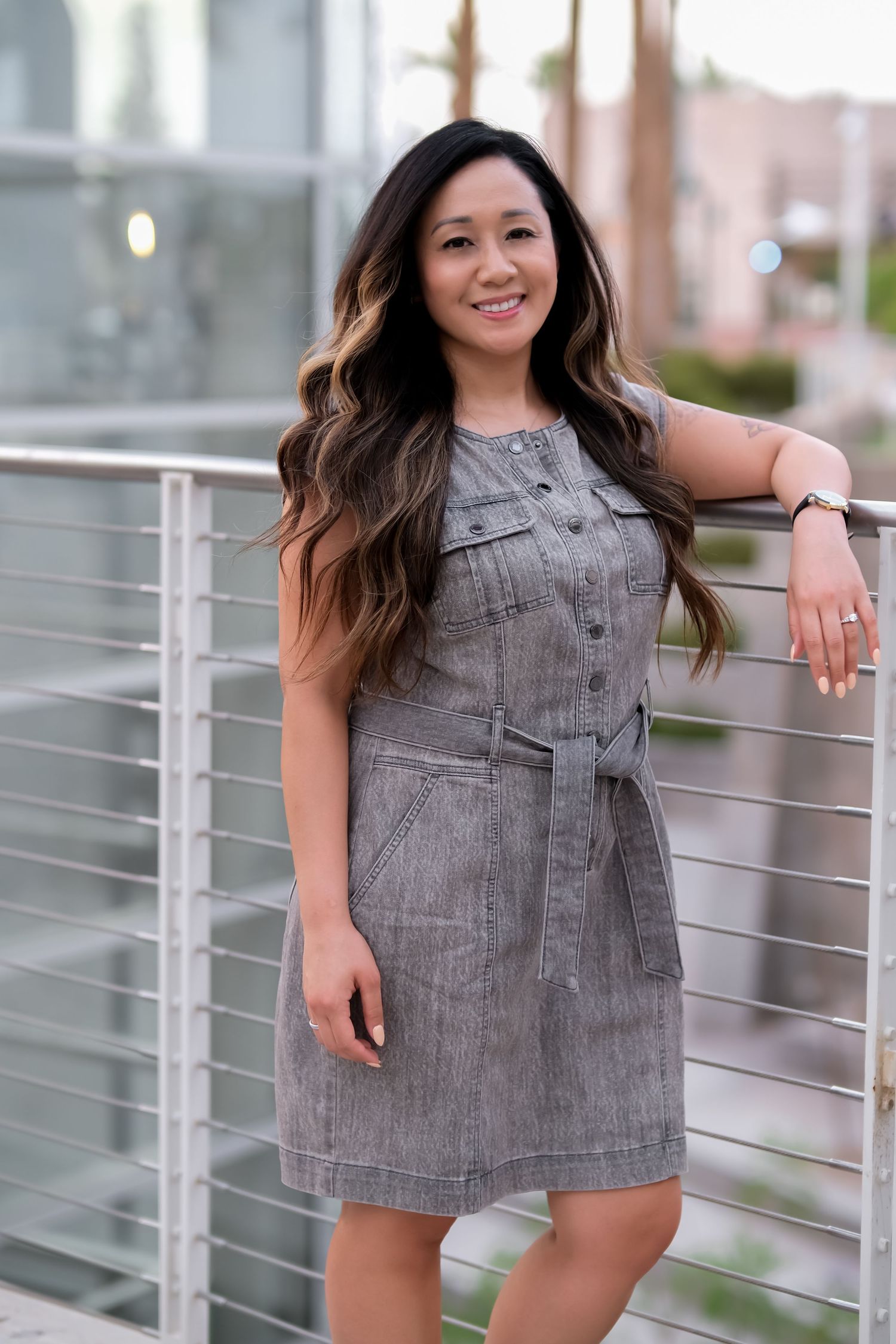 Woman in gray denim dress poses casually while leaning against metal railing outdoors.