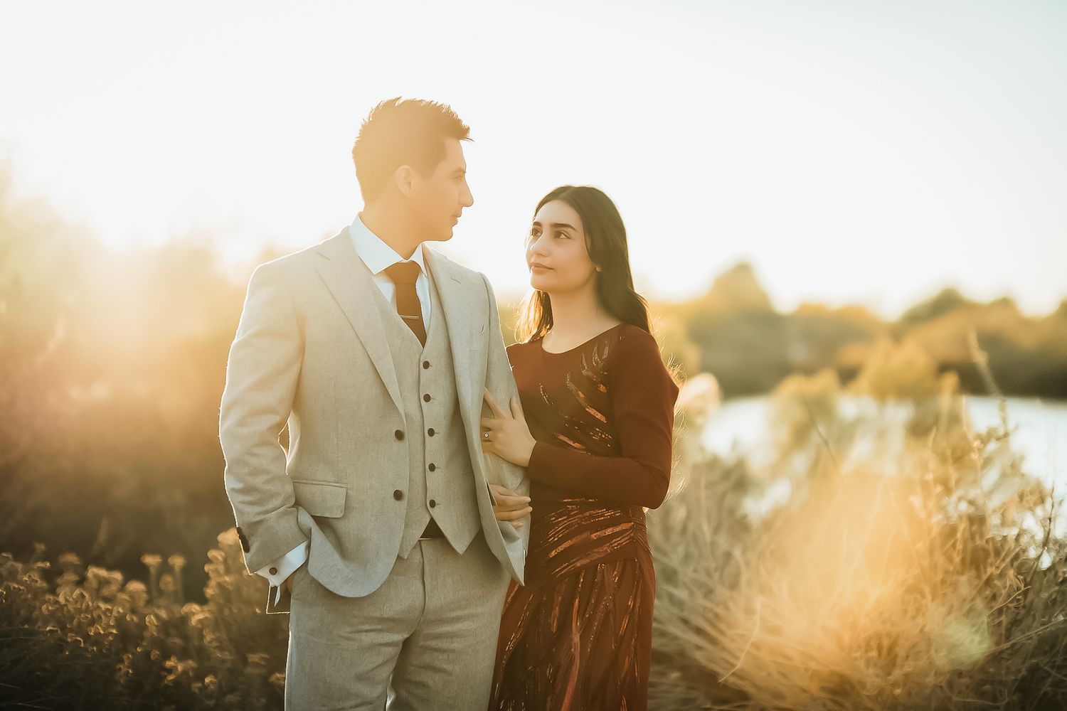 A couple in formal wear poses against golden sunlight near a waterfront.