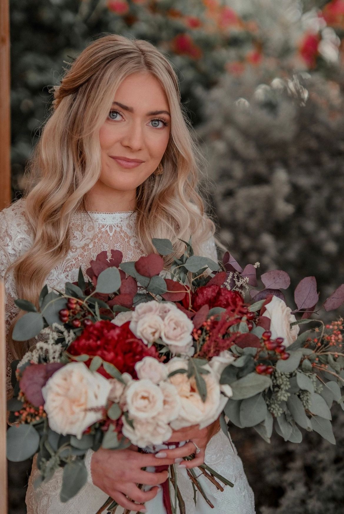 Bride holds stunning bouquet of burgundy and blush roses with eucalyptus against autumn background.