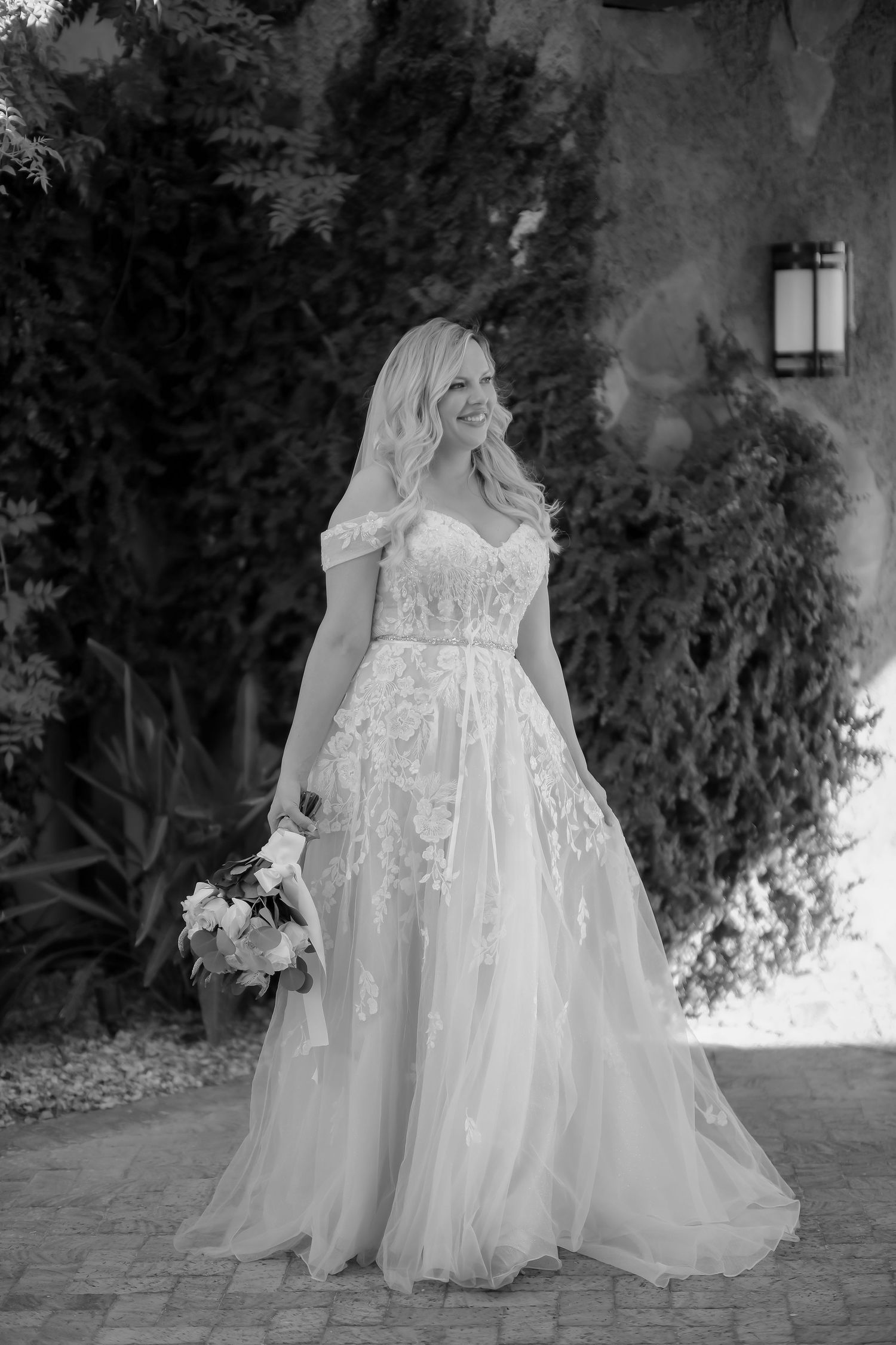 Black and white portrait of bride in lace gown holding bouquet on stone pathway.