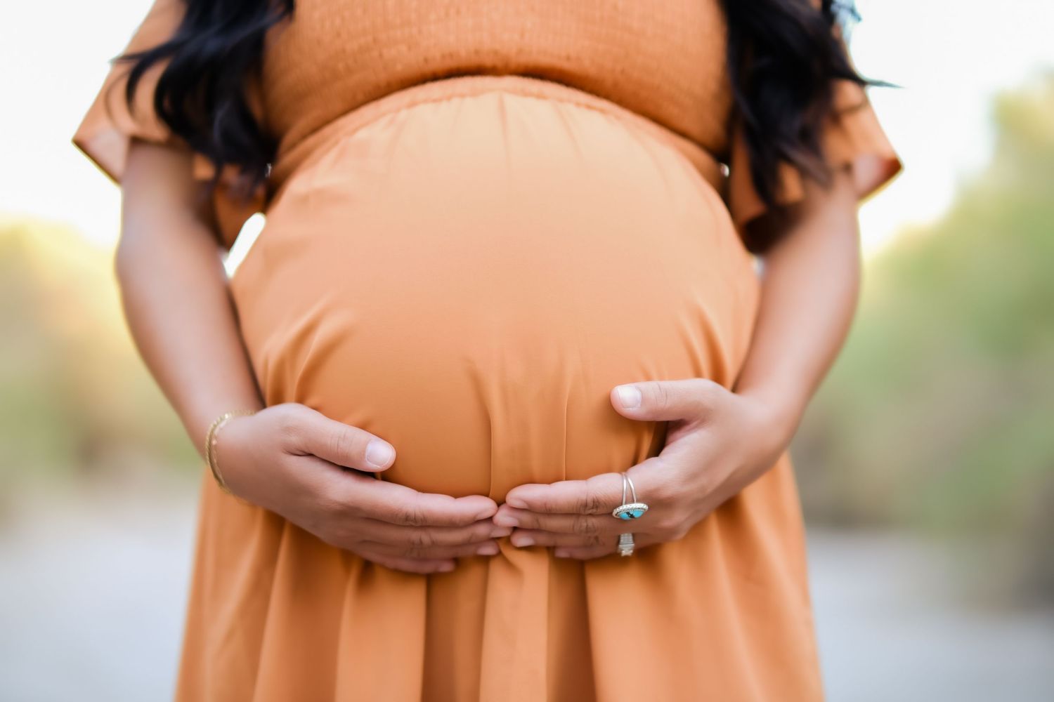 Close-up of hands cradling a pregnant belly in an orange maternity dress with an engagement ring.