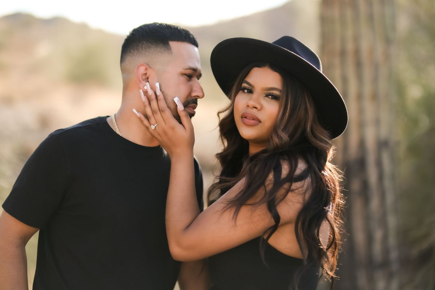 A stylish couple shares an intimate moment outdoors wearing black attire with a wide-brimmed hat.