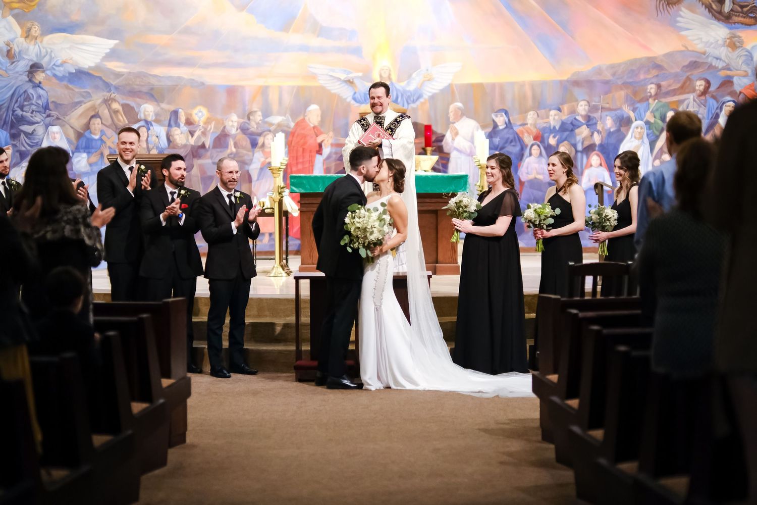 Bride and groom share first kiss at church altar surrounded by wedding party and applauding guests.