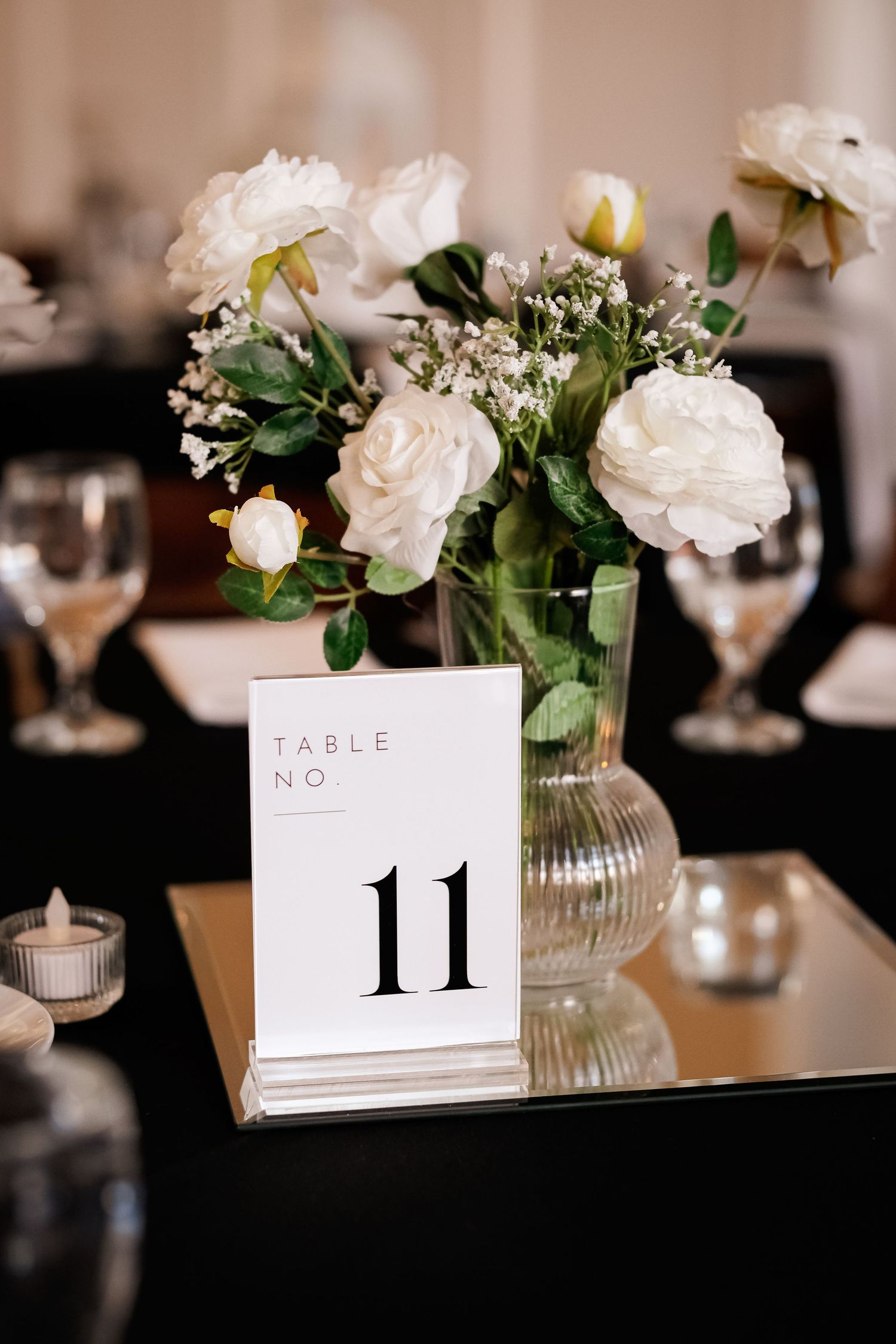 Elegant wedding table 11 centerpiece with white roses, greenery in glass vase on mirrored surface with candles and wine glasses.