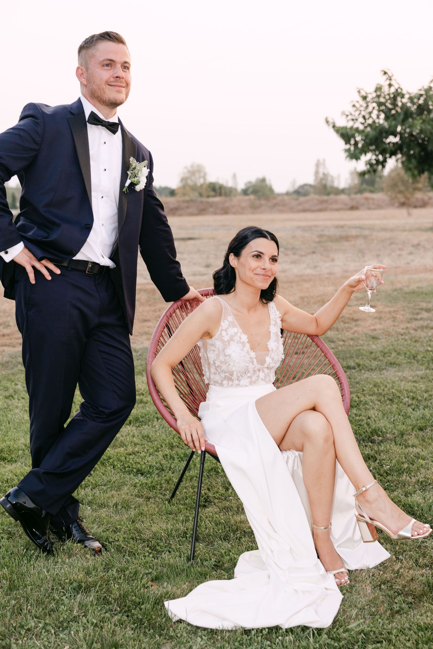 A bride in a lace wedding gown and groom in dark tuxedo sit playfully on grass during an outdoor photo session.