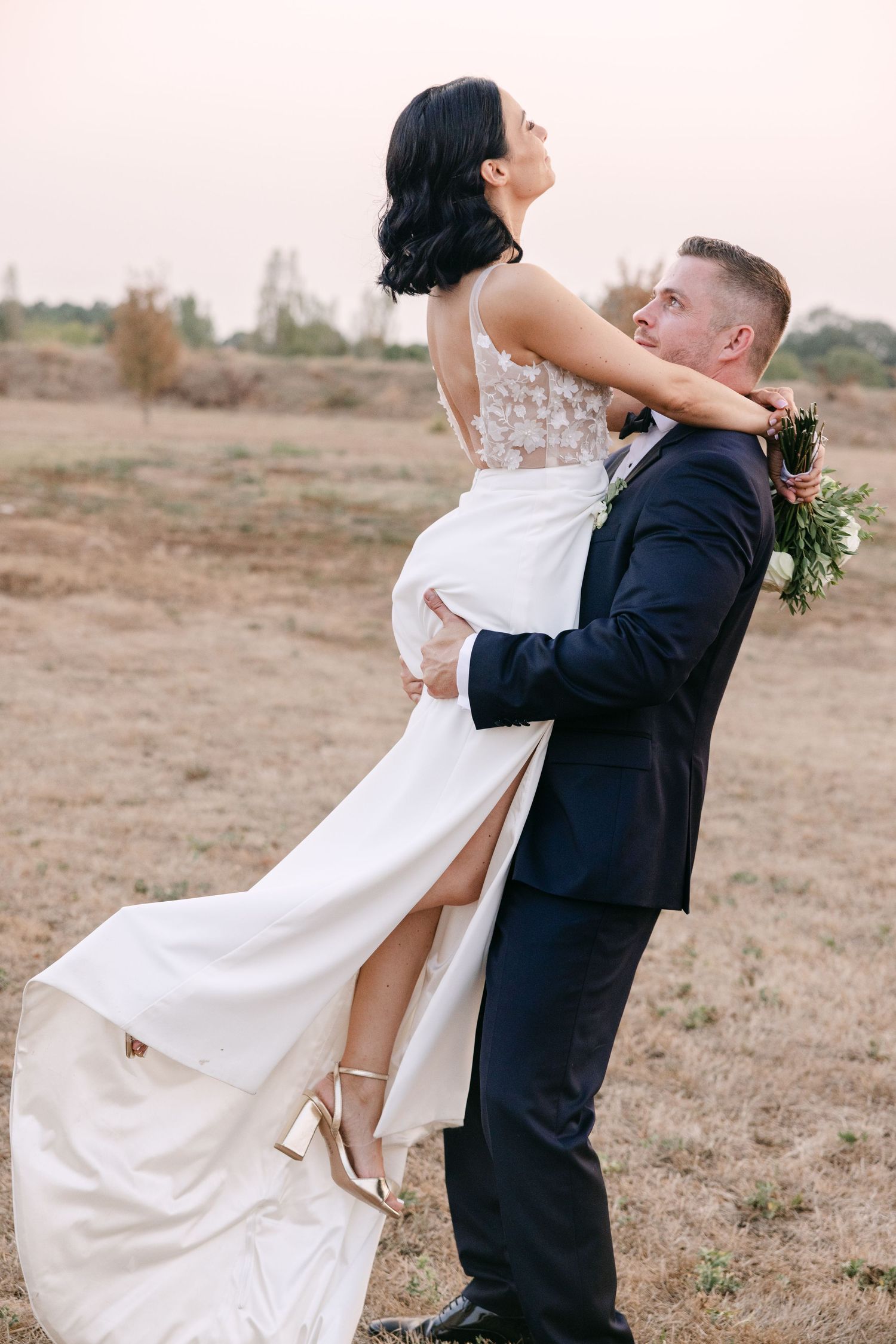 A joyful couple in wedding attire share playful dance moves together in an outdoor field setting.