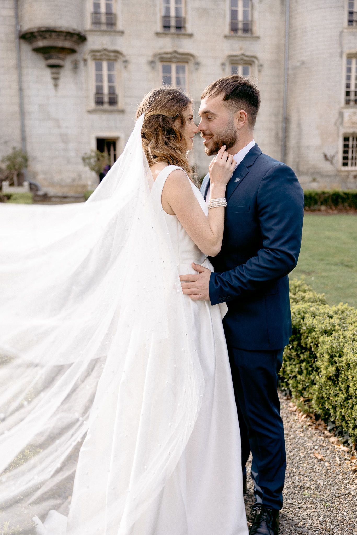 Couple sharing intimate moment outside historic chateau with flowing wedding veil.