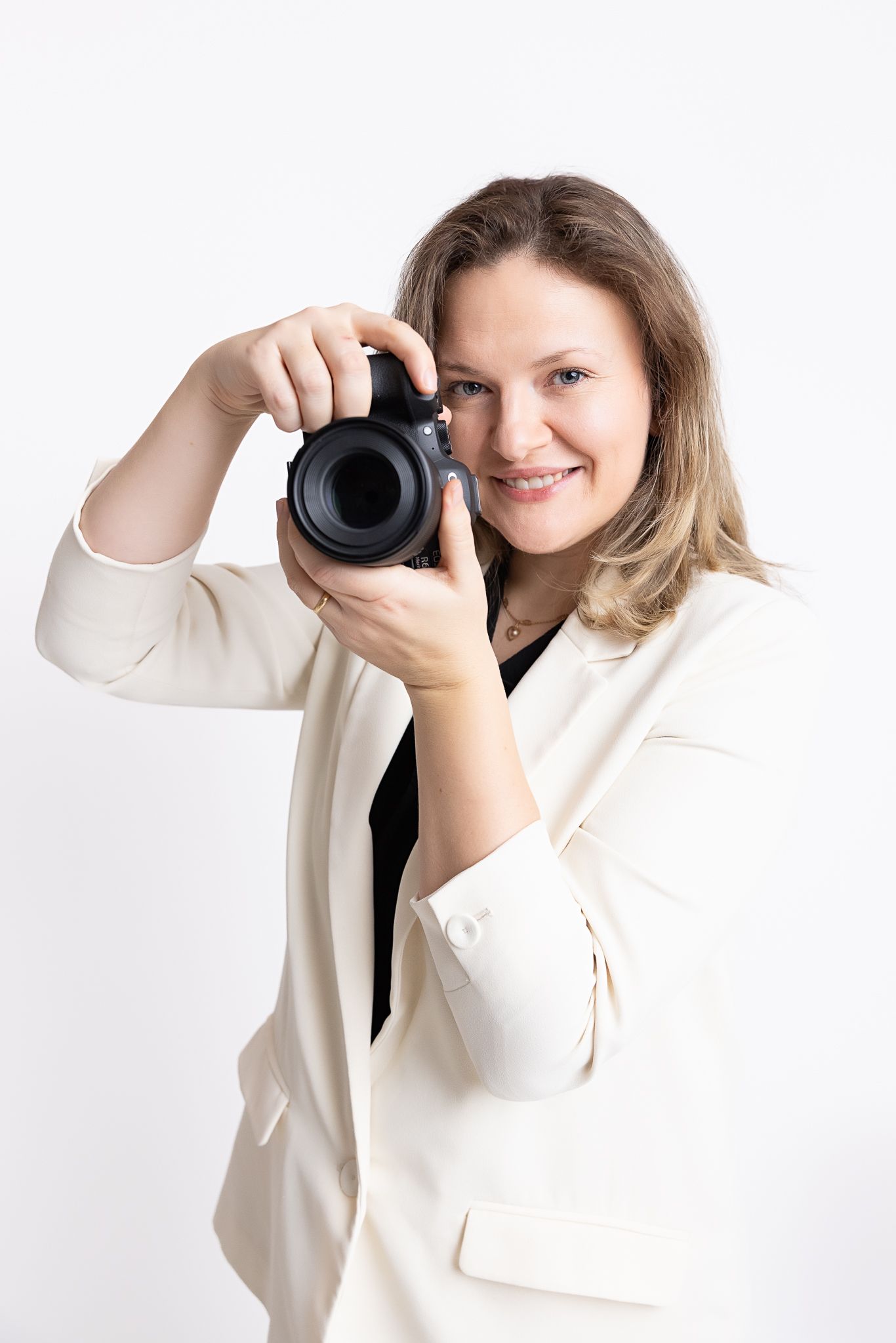 Professional photographer in white blazer holding DSLR camera while smiling against light background.