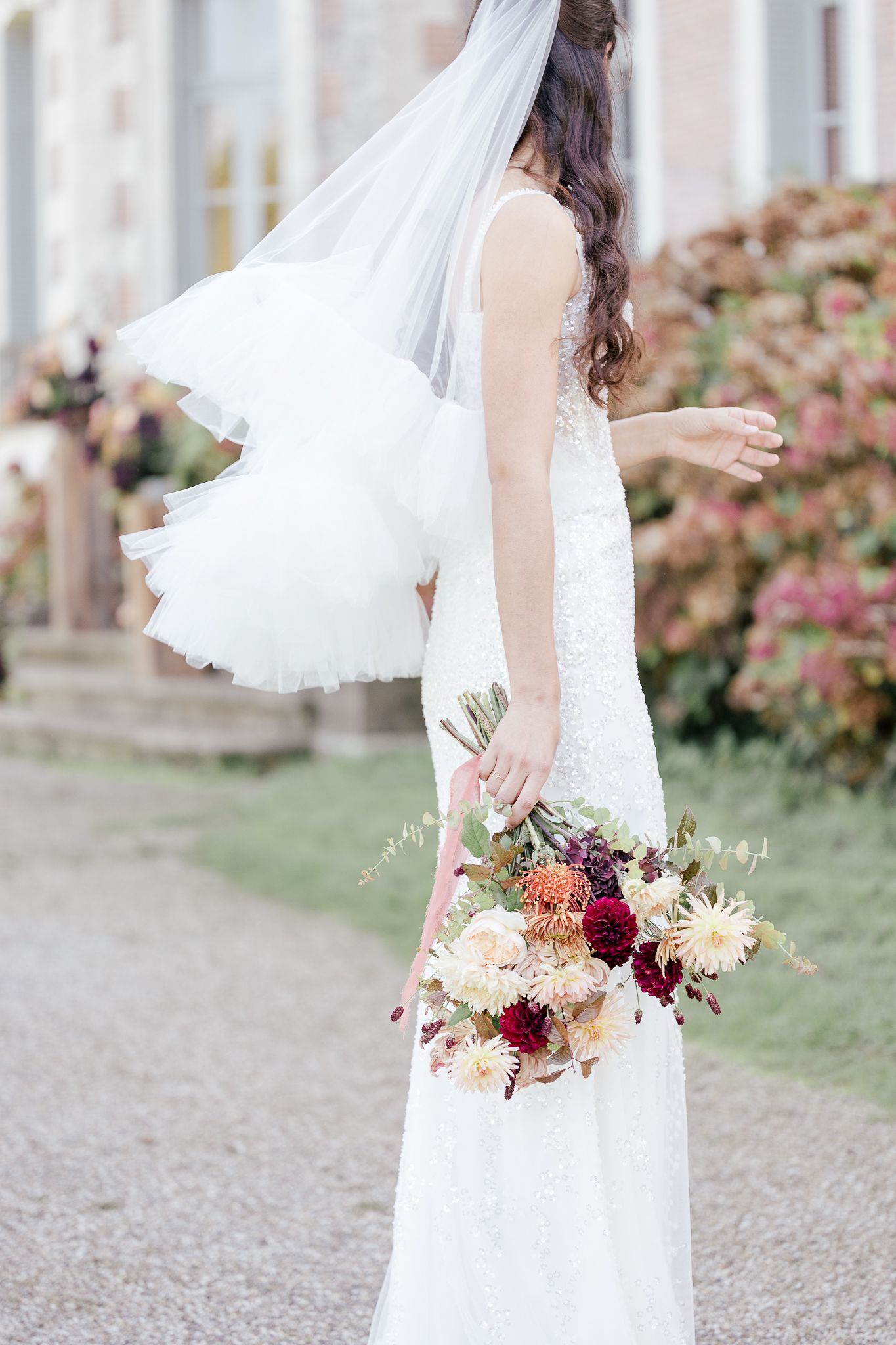 Side view of bride in white lace dress and veil holding romantic burgundy and blush wedding bouquet.