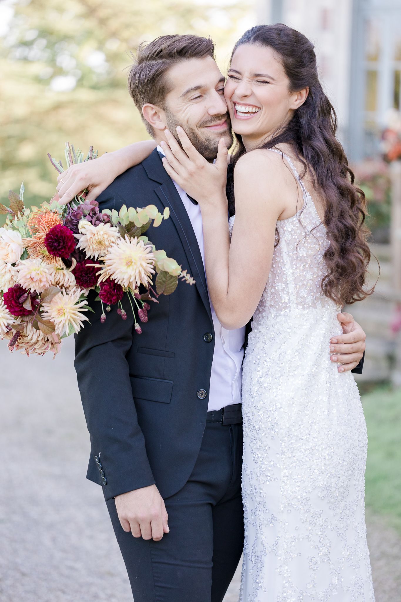A joyful wedding couple shares a tender moment while holding a vibrant autumn bouquet with burgundy and blush flowers.