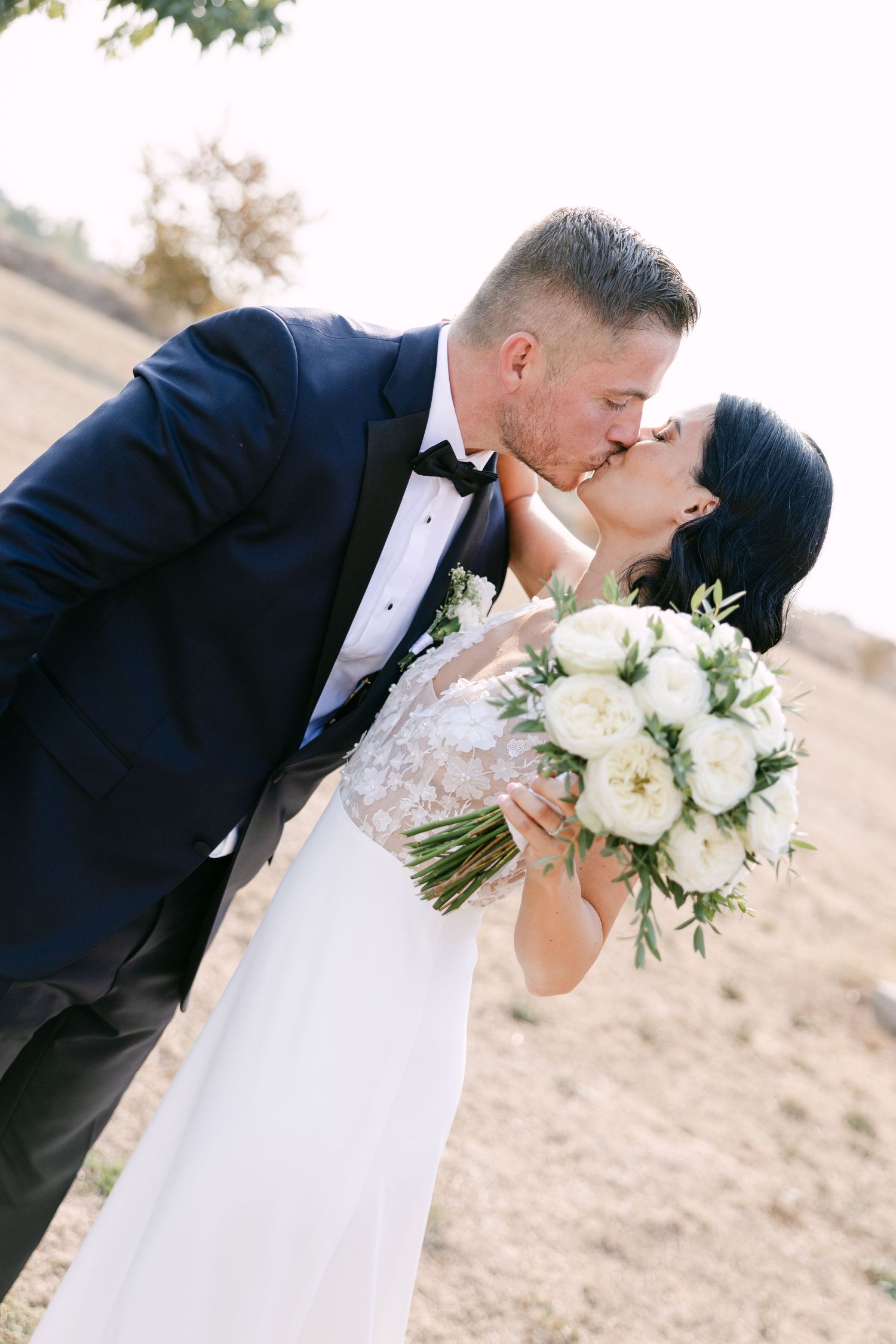 Elegant newlyweds share a romantic kiss while bride holds stunning white peony bouquet at outdoor wedding.