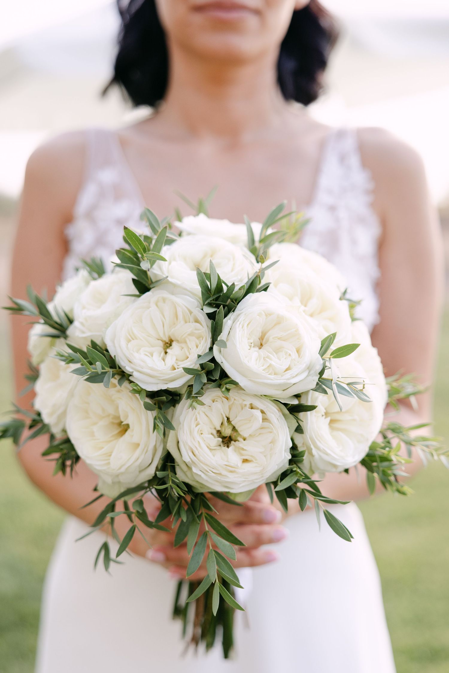 Four elegant white rose bouquets with delicate greenery held against white dresses in an outdoor setting.