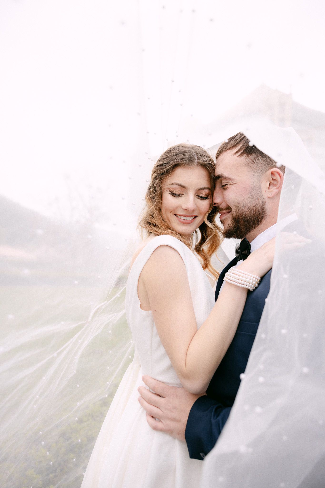 Dreamy outdoor wedding portrait with flowing veil against misty landscape.
