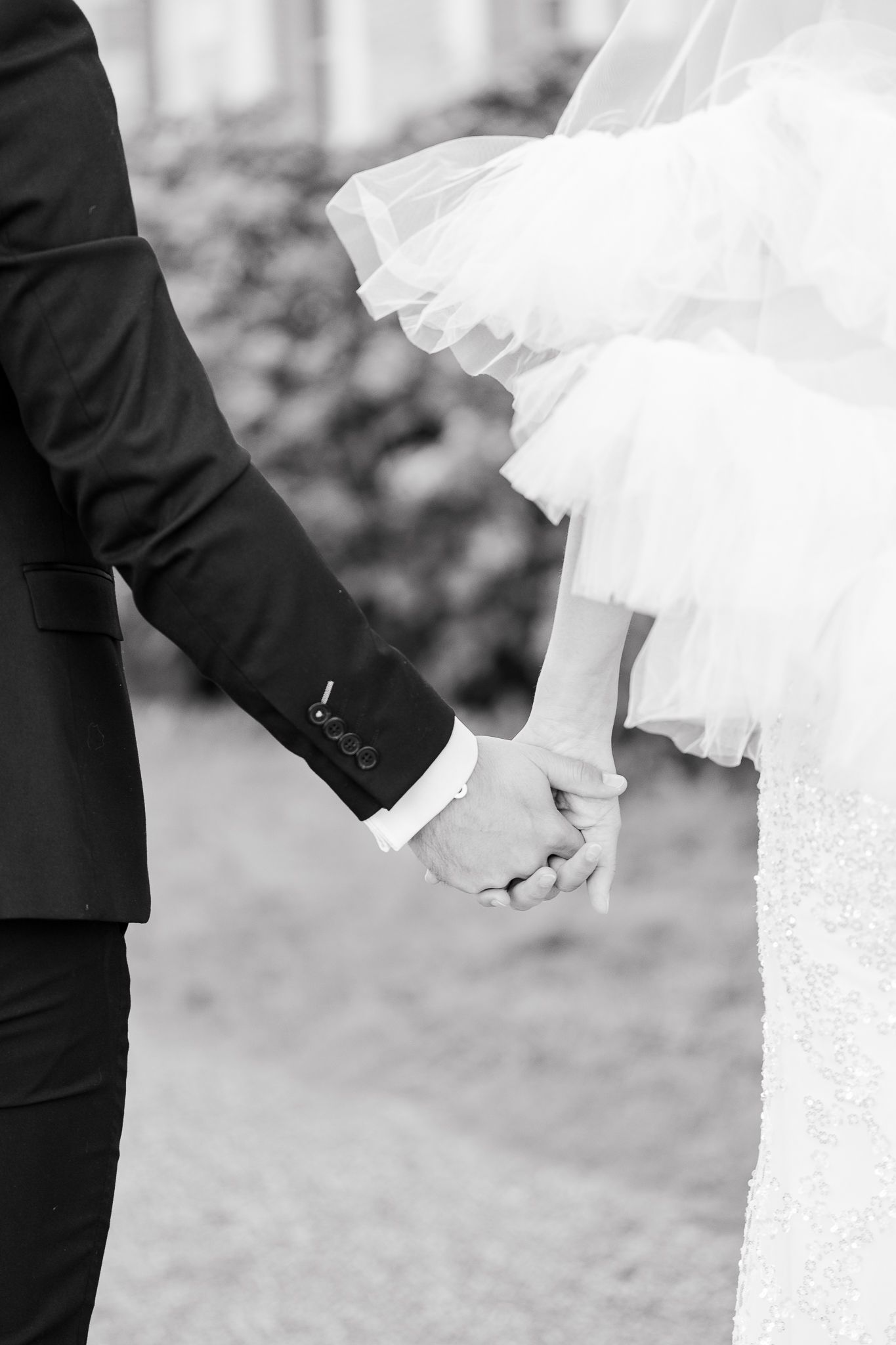 Black and white romantic image of newlywed couple holding hands with bride in flowing white dress and groom in suit.