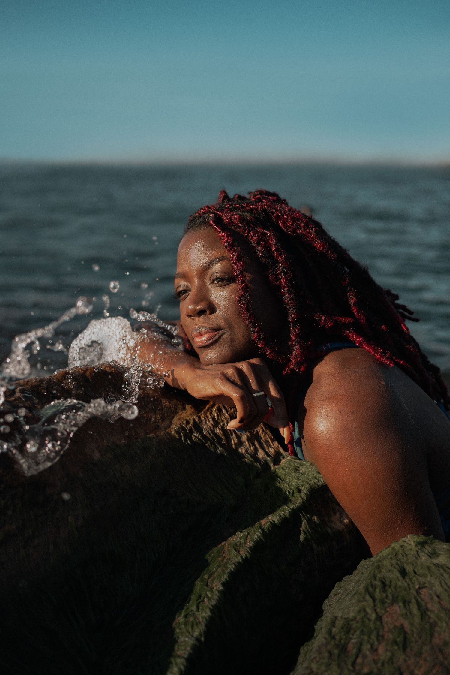 Ocean waves splash around a figure resting against a mossy rock during a beach photoshoot.