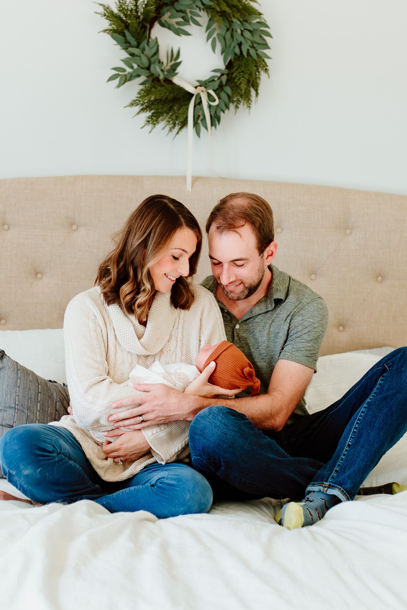 Couple cuddles with newborn baby on bed beneath holiday wreath. Charleston, South Carolina. Salt and Oak Photography.