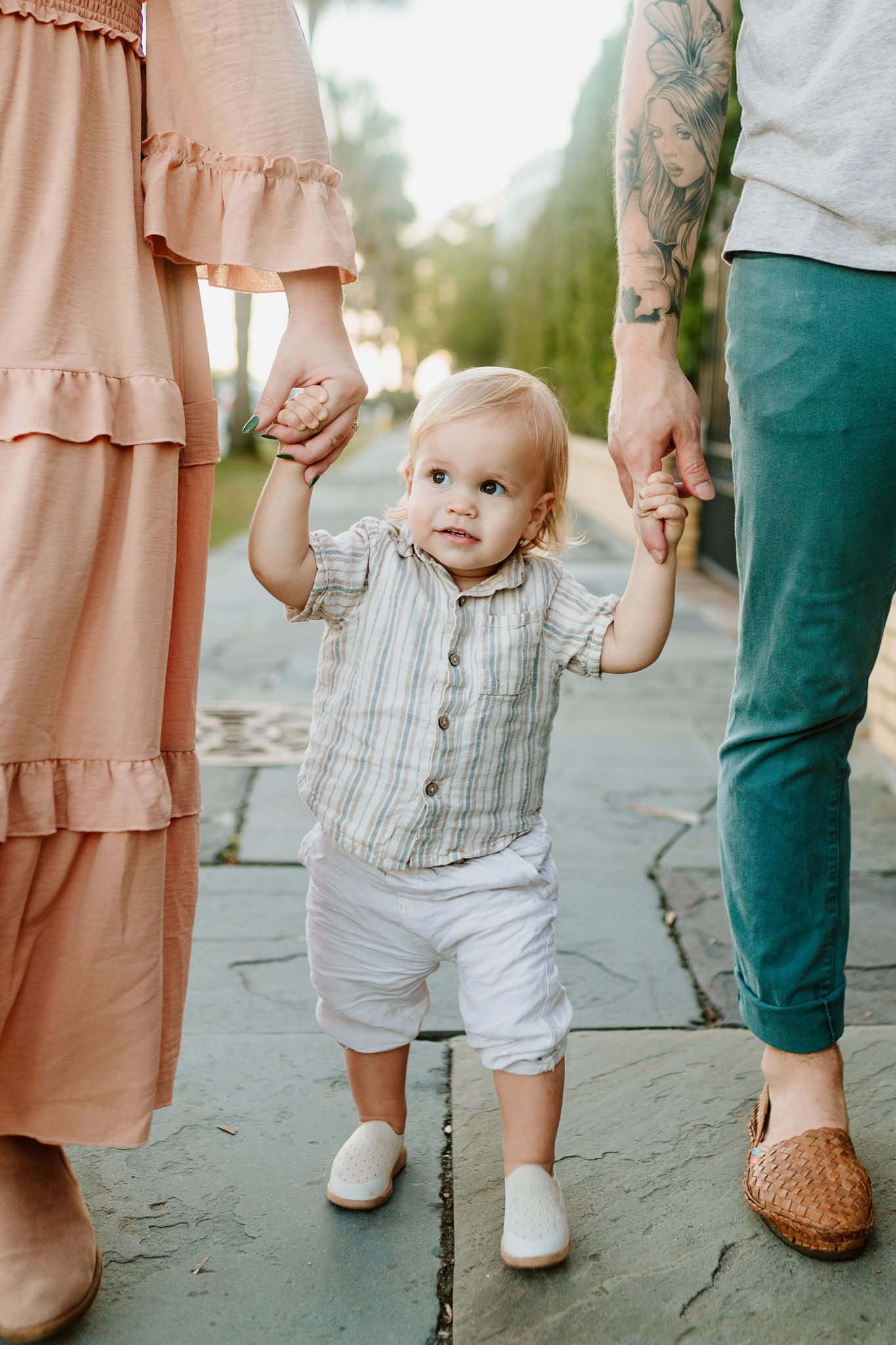 A toddler takes first steps while holding parents' hands on a stone pathway. Charleston, South Carolina. Salt and Oak Photography.