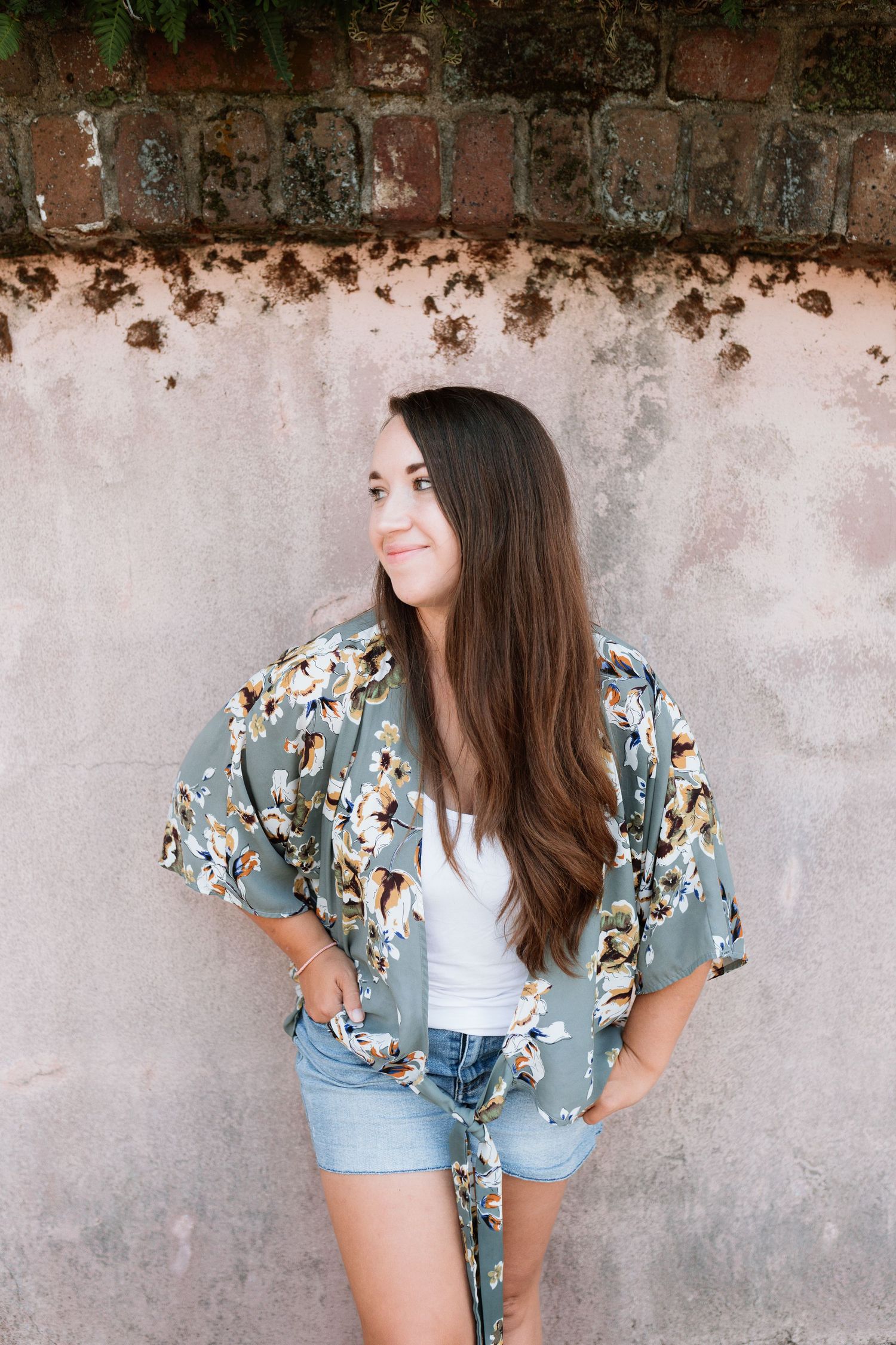Woman in floral print kimono and denim shorts poses against weathered concrete wall.