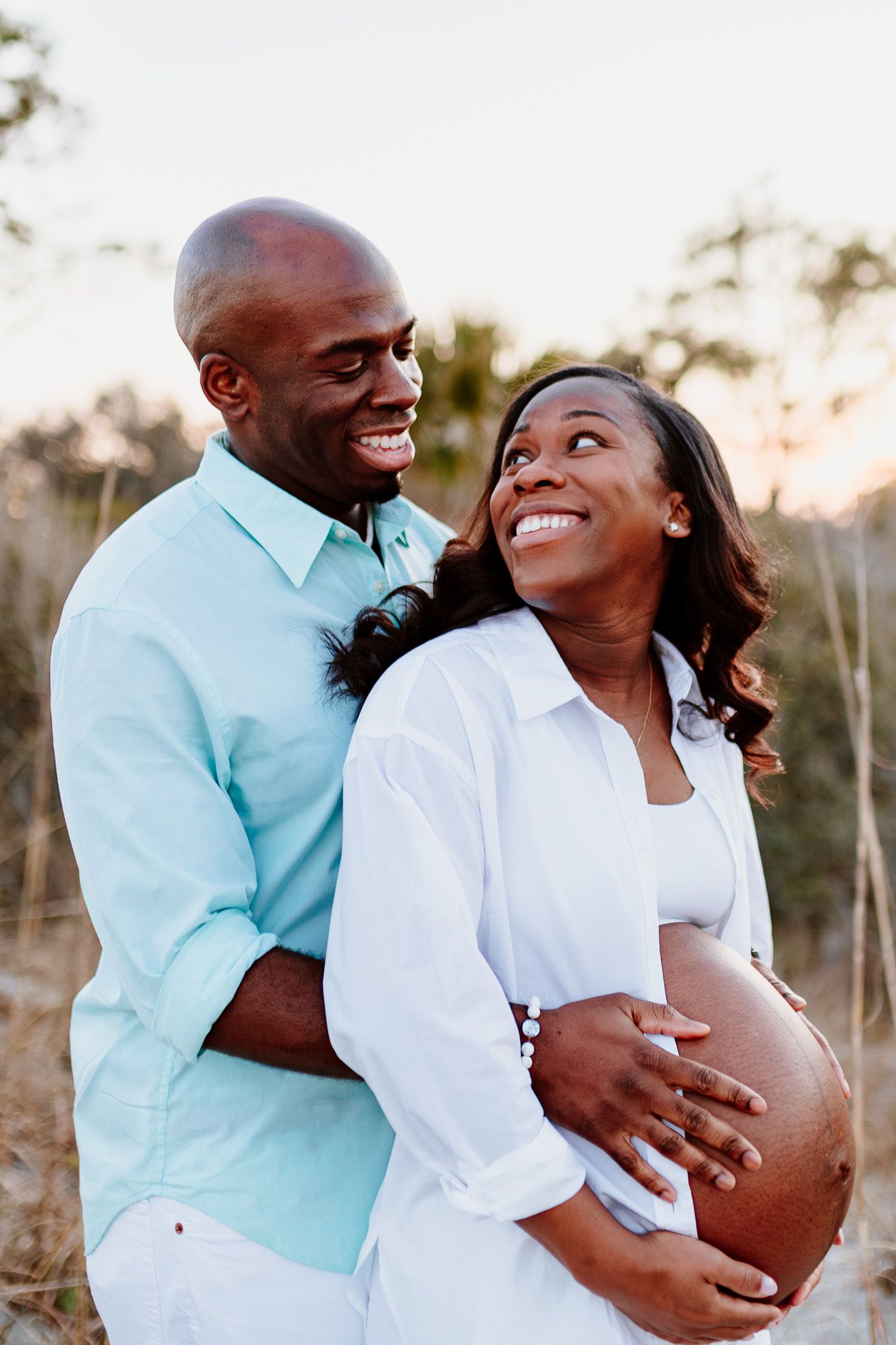 A loving couple embraces outdoors during sunset as they share a joyful pregnancy. Charleston, South Carolina. Salt and Oak Photography.