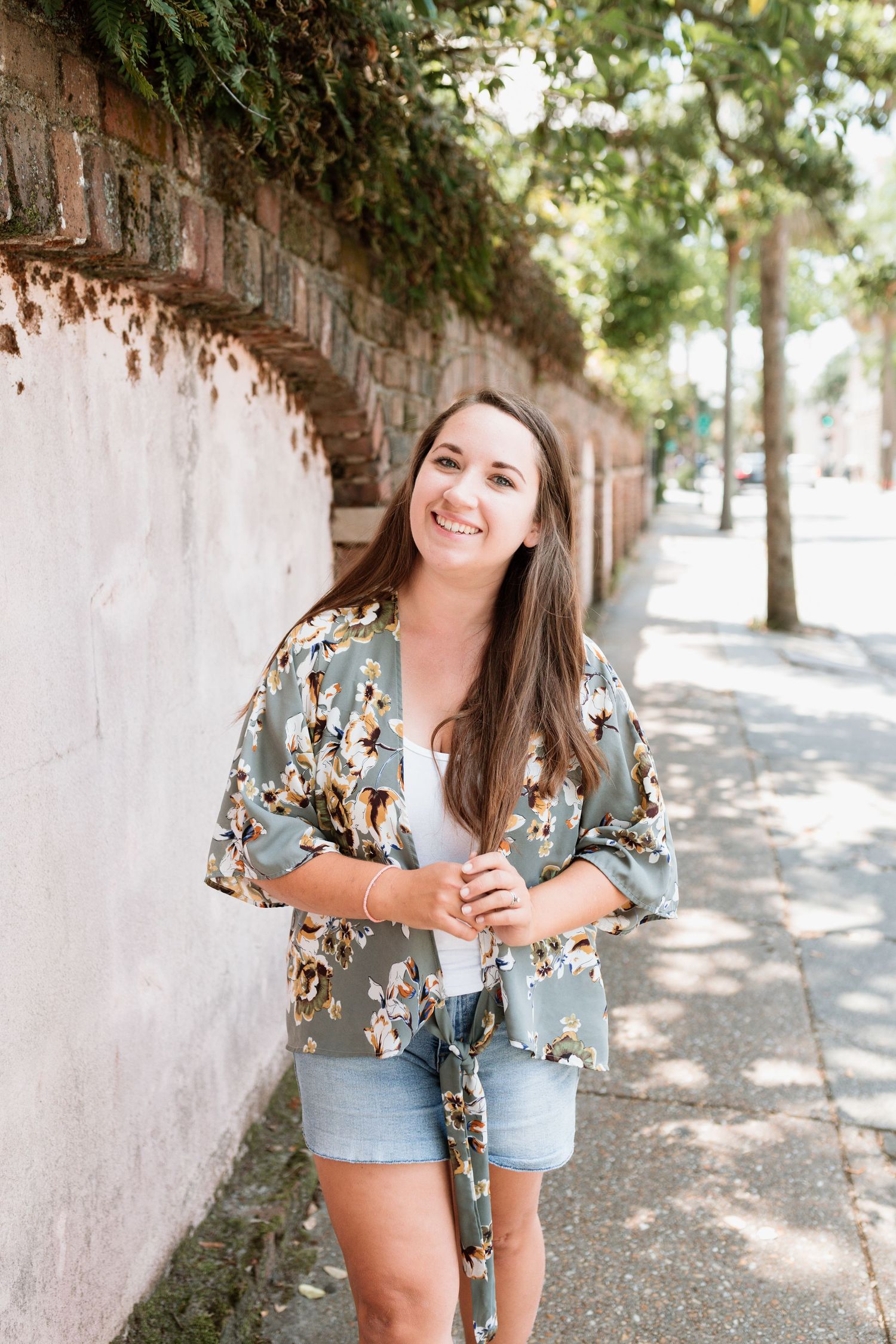 A person in a floral kimono-style top and denim shorts stands smiling along a tree-lined path beside a brick wall.