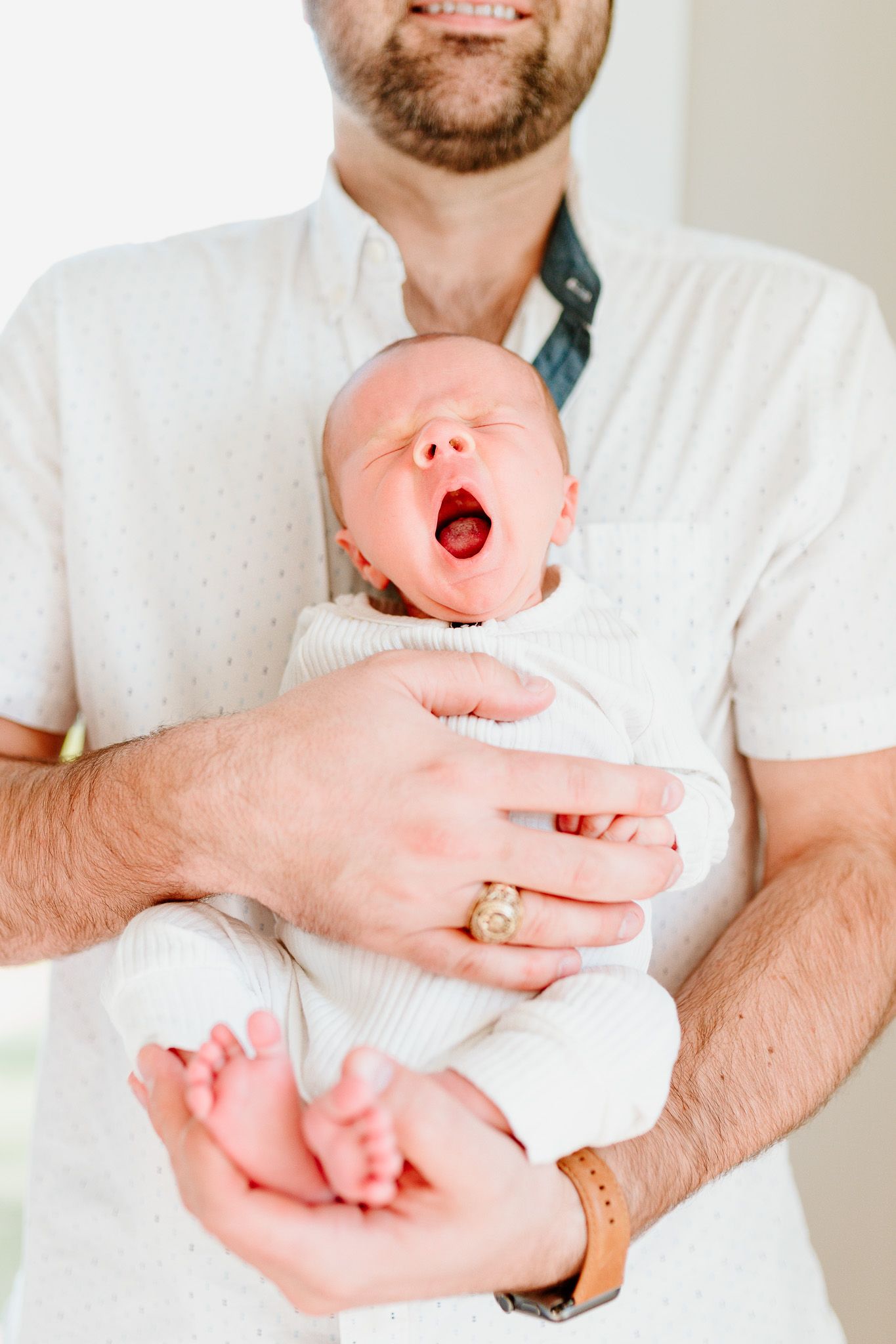 A yawning newborn baby being held in white onesie against white background. Charleston, South Carolina. Salt and Oak Photography.